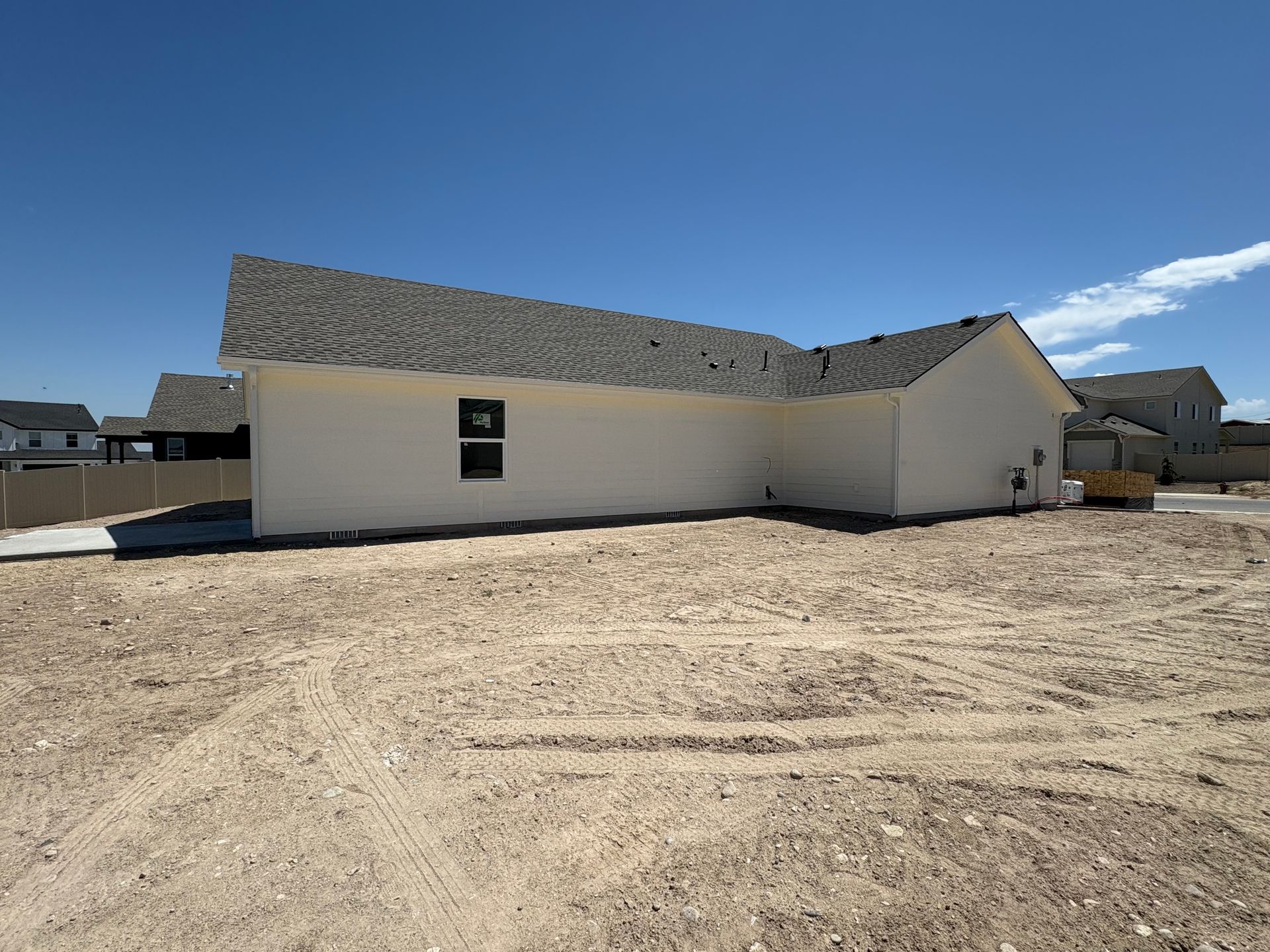 A newly constructed, single-story house with light-colored siding and a gray roof sits on a lot of dirt under a blue sky.