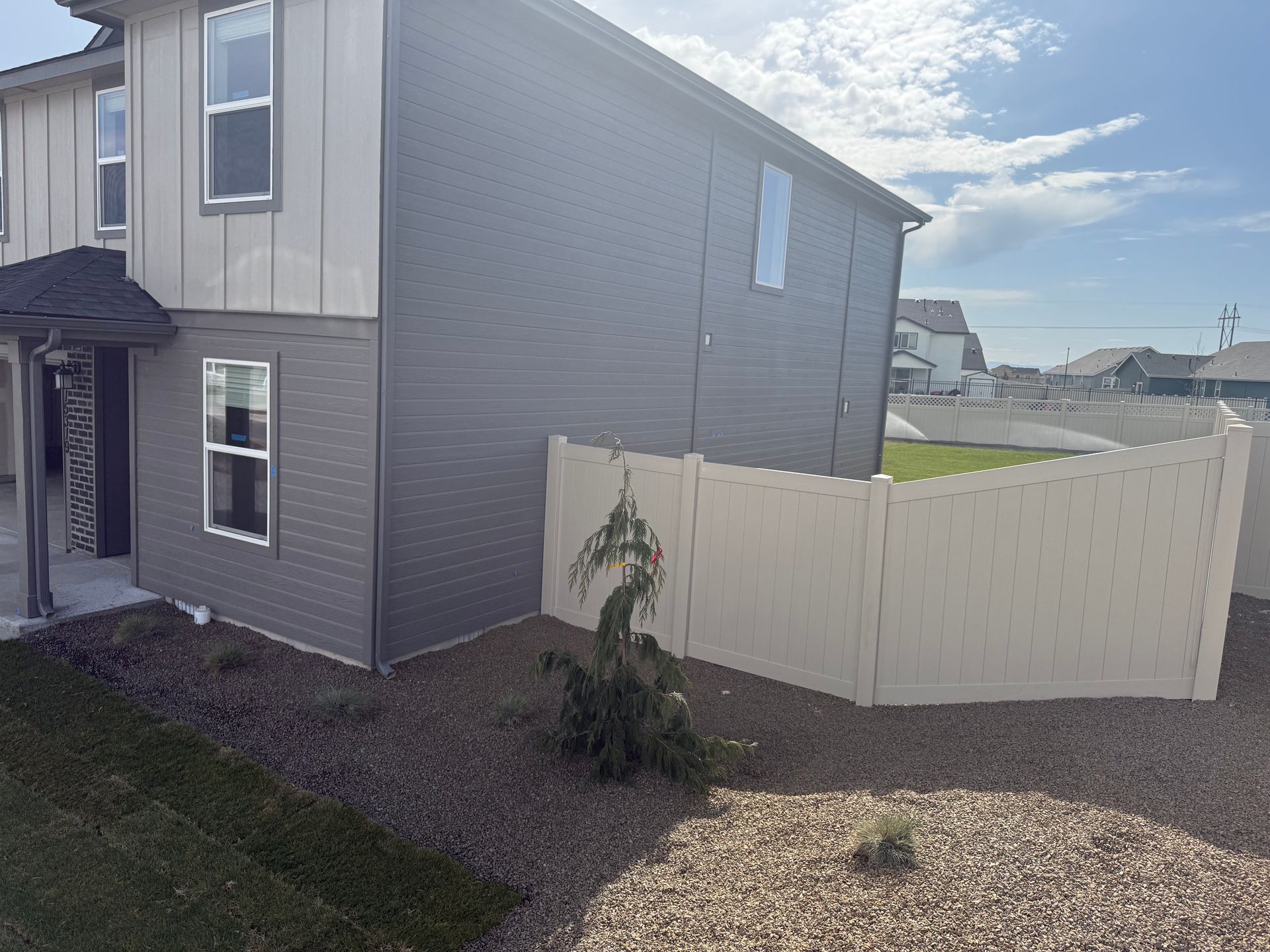 Side view of a two-story house with light and dark gray siding and a beige fence, sunny day.