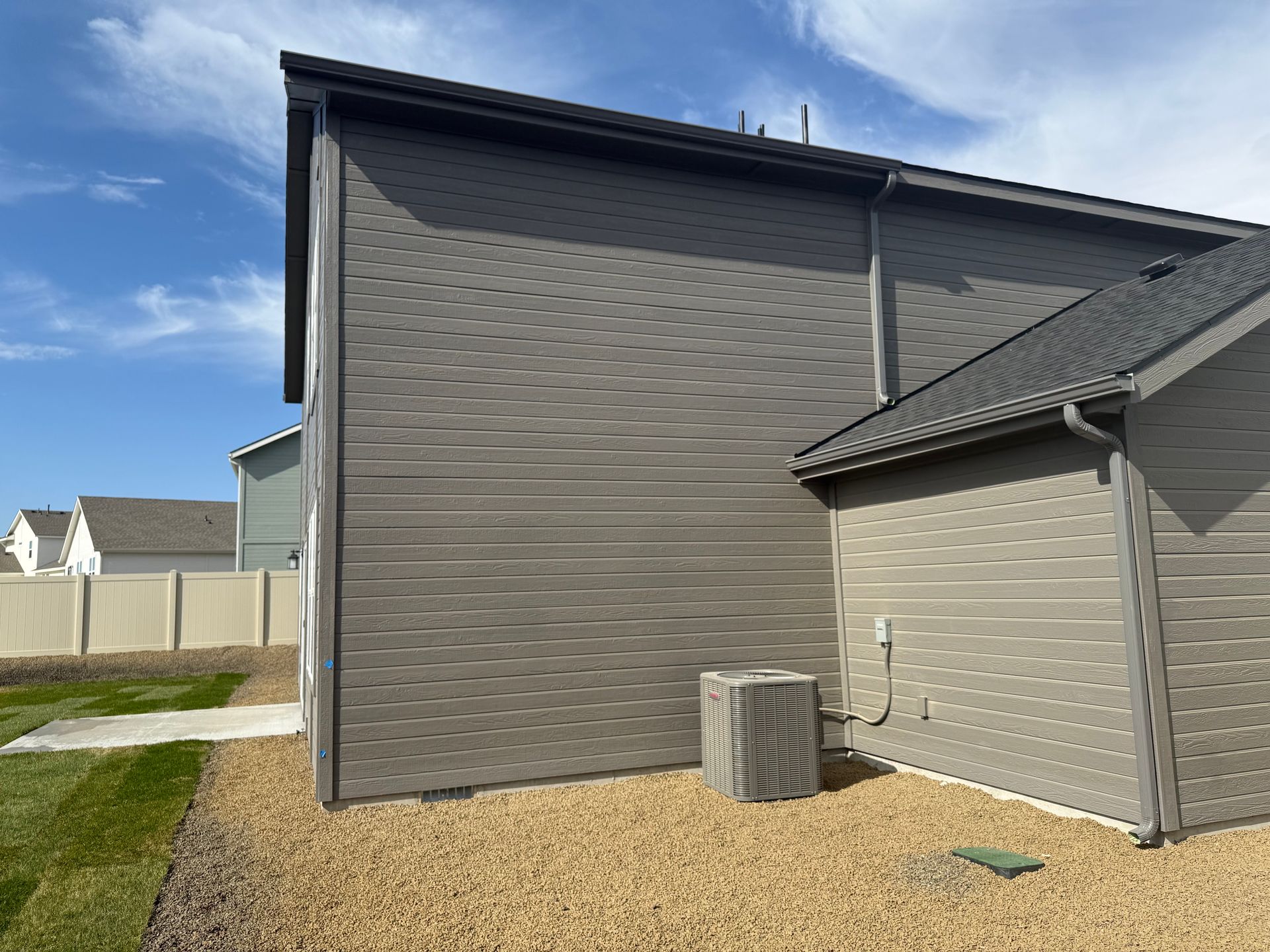 Gray-sided house exterior with air conditioning unit on a gravel bed; blue sky.