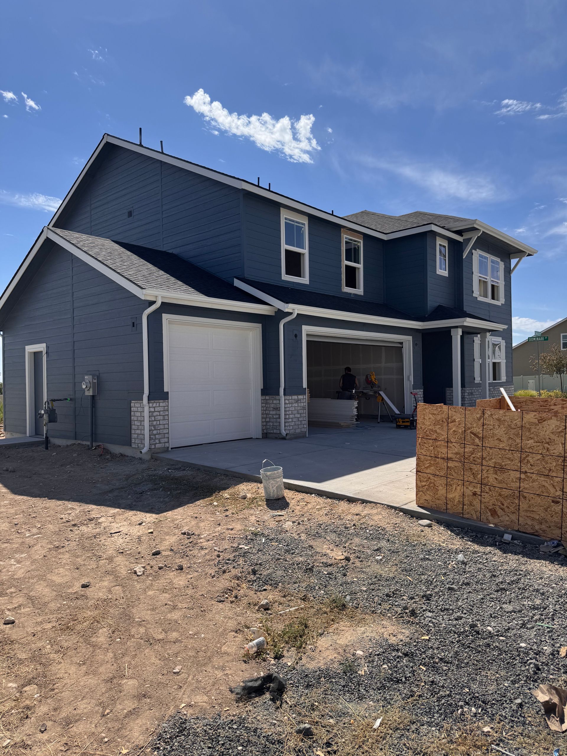 Two-story blue house with white trim and a two-car garage under construction, set on gravel, with a partly cloudy sky.