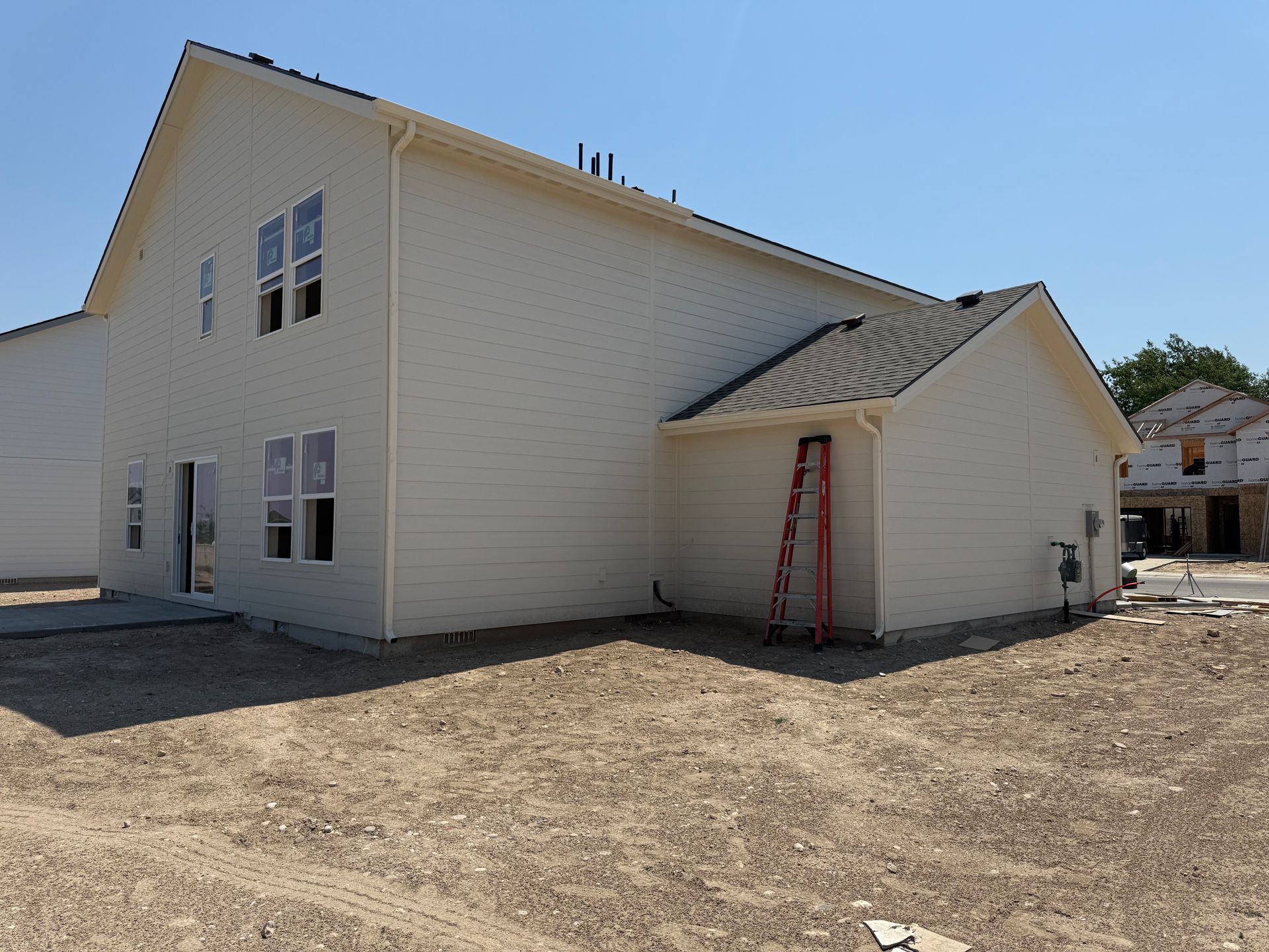 Side view of a light-colored two-story house under construction, with a red ladder and bare earth.
