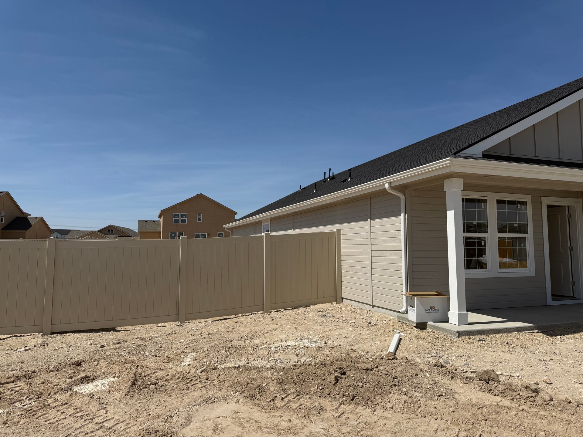 Beige fence bordering a house with beige siding and a black roof on a sunny day.