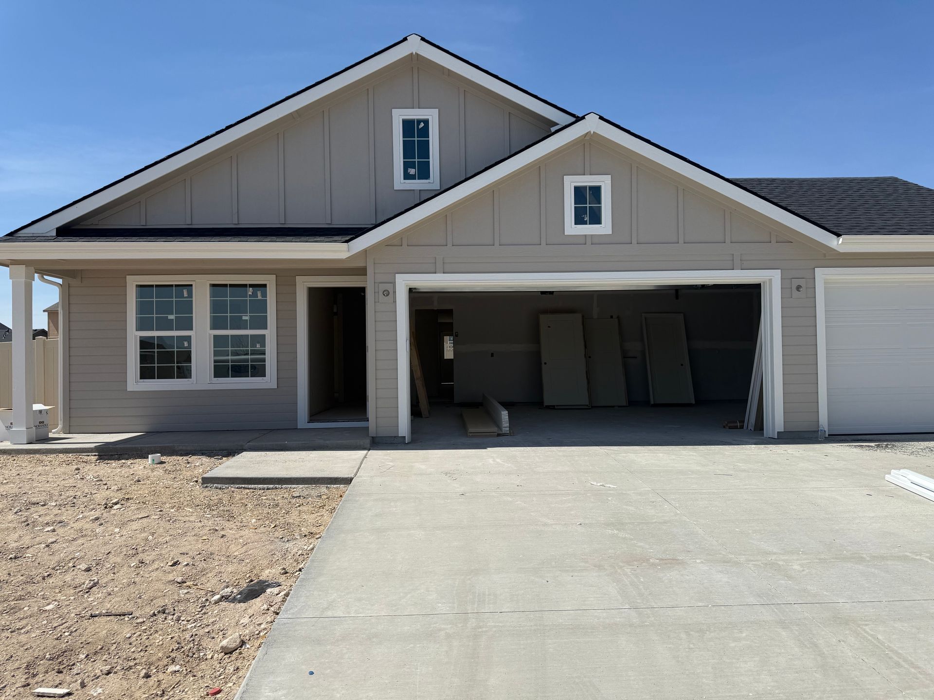 Tan house with attached garage, unfinished construction. Concrete driveway and bare earth in front.