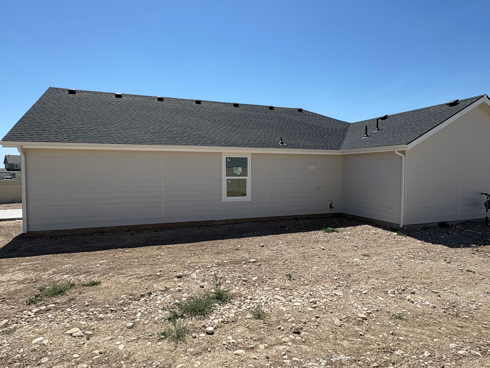 Tan house exterior with gray roof and a window on gravel ground under a blue sky.
