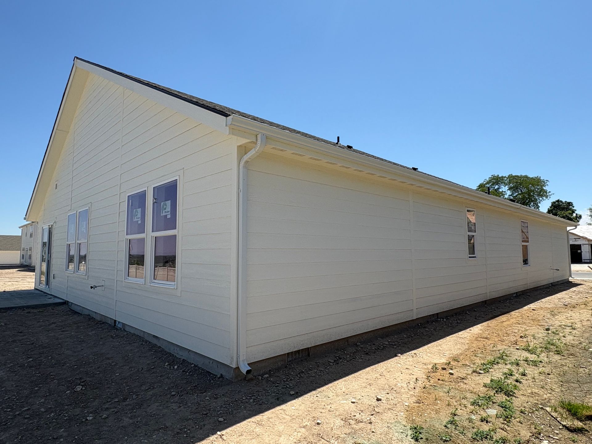 Side view of a new, beige house under a clear blue sky; windows covered with plastic, gravel ground.