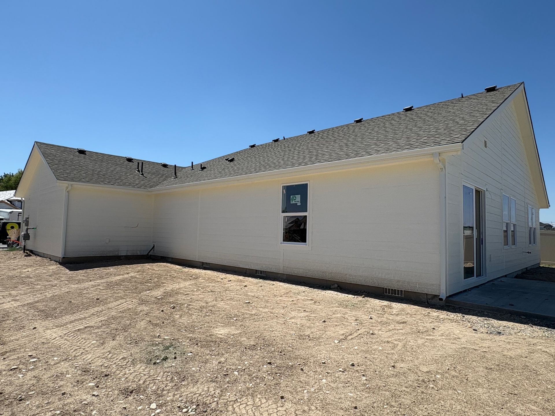Side view of a light-colored house with a dark roof on a sunny day; dirt lot in front.