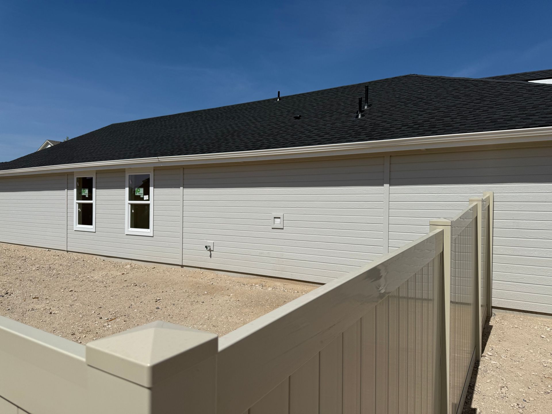Side of a light gray house with black roof, two windows, and beige fence on a gravel lot under a blue sky.