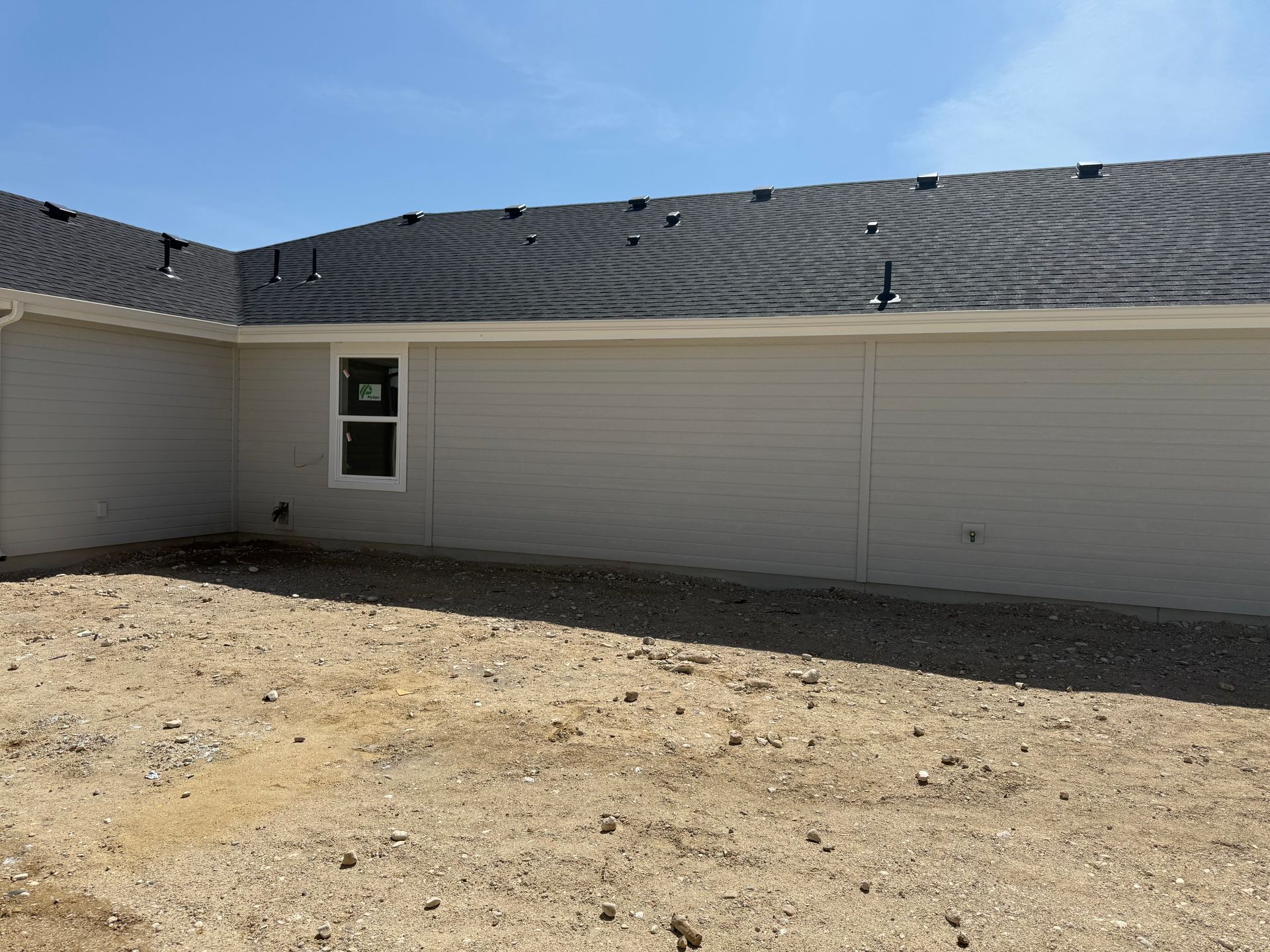 Beige house exterior with a window, dark roof, and dirt yard under a blue sky.