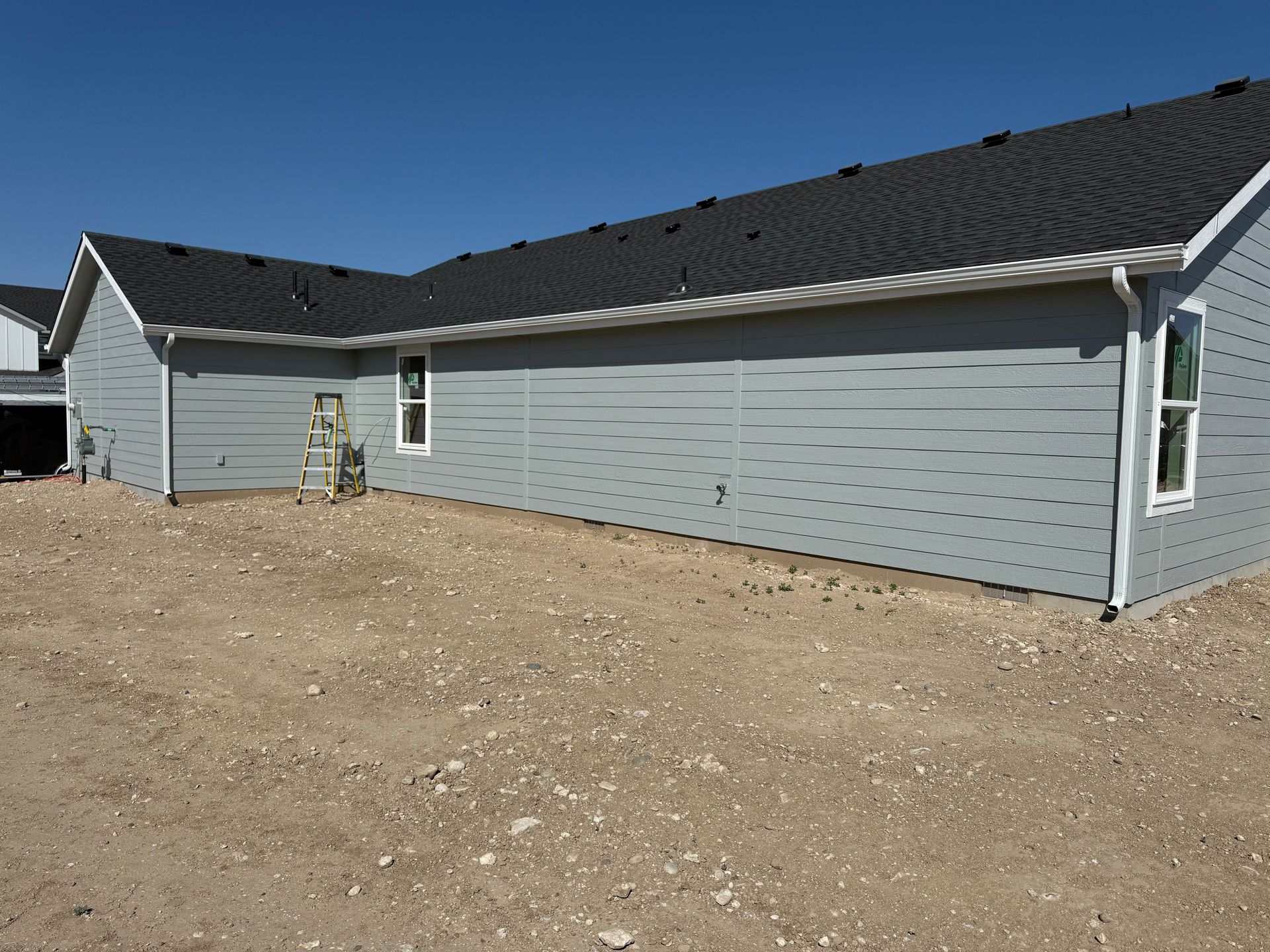 Gray house with white trim, black roof, and gravel ground. Clear, blue sky.