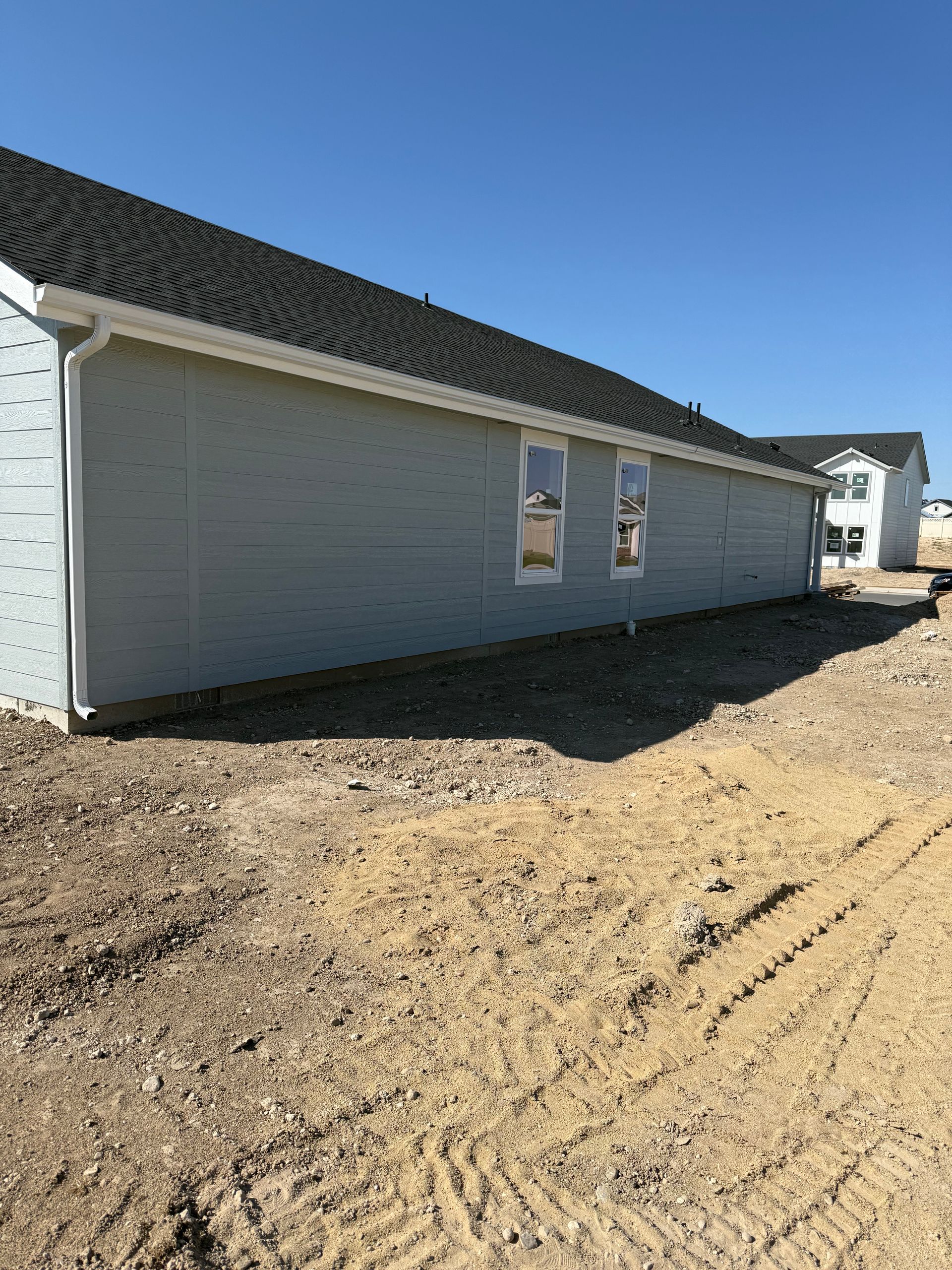 Side of a light blue house with black roof and white window frames, on a dirt lot, under a clear blue sky.