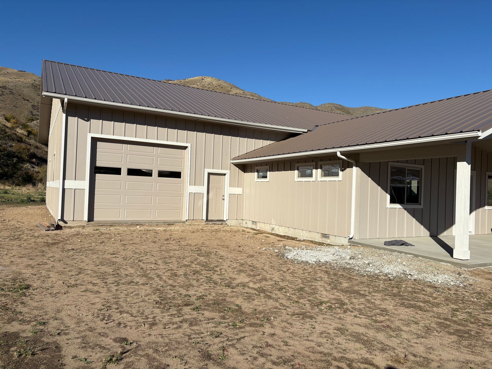 Beige building with a garage door, side door, and windows, brown roof, set against a mountainous landscape.