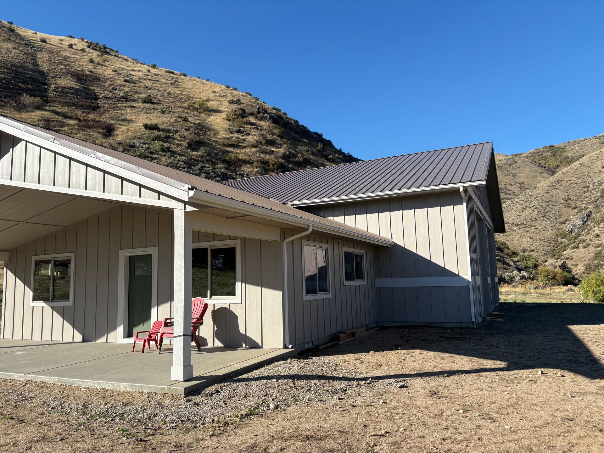 Beige house with a metal roof against a mountain backdrop under a blue sky.