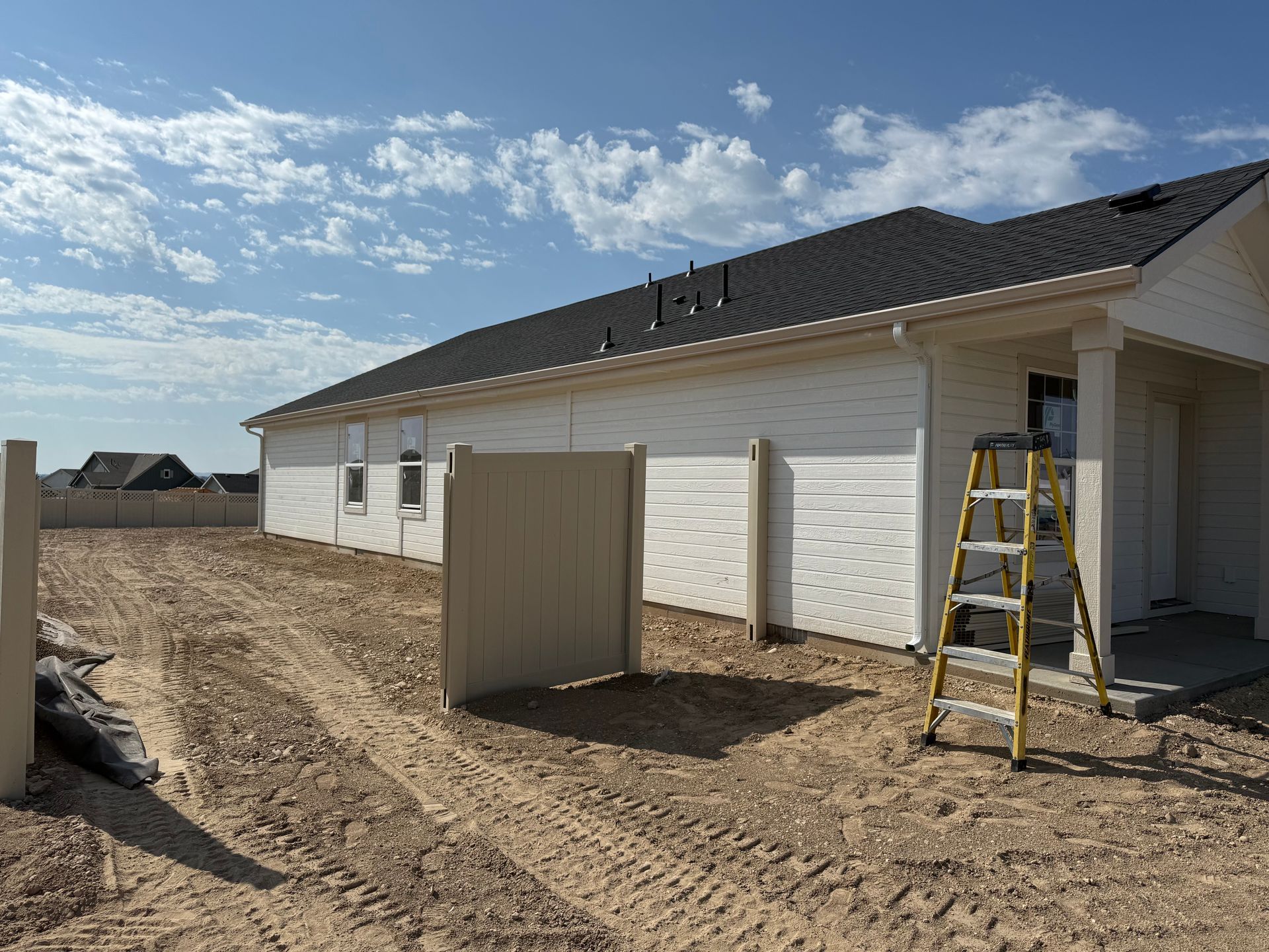Exterior view of a house under construction with a tan fence, a ladder, and a blue sky.
