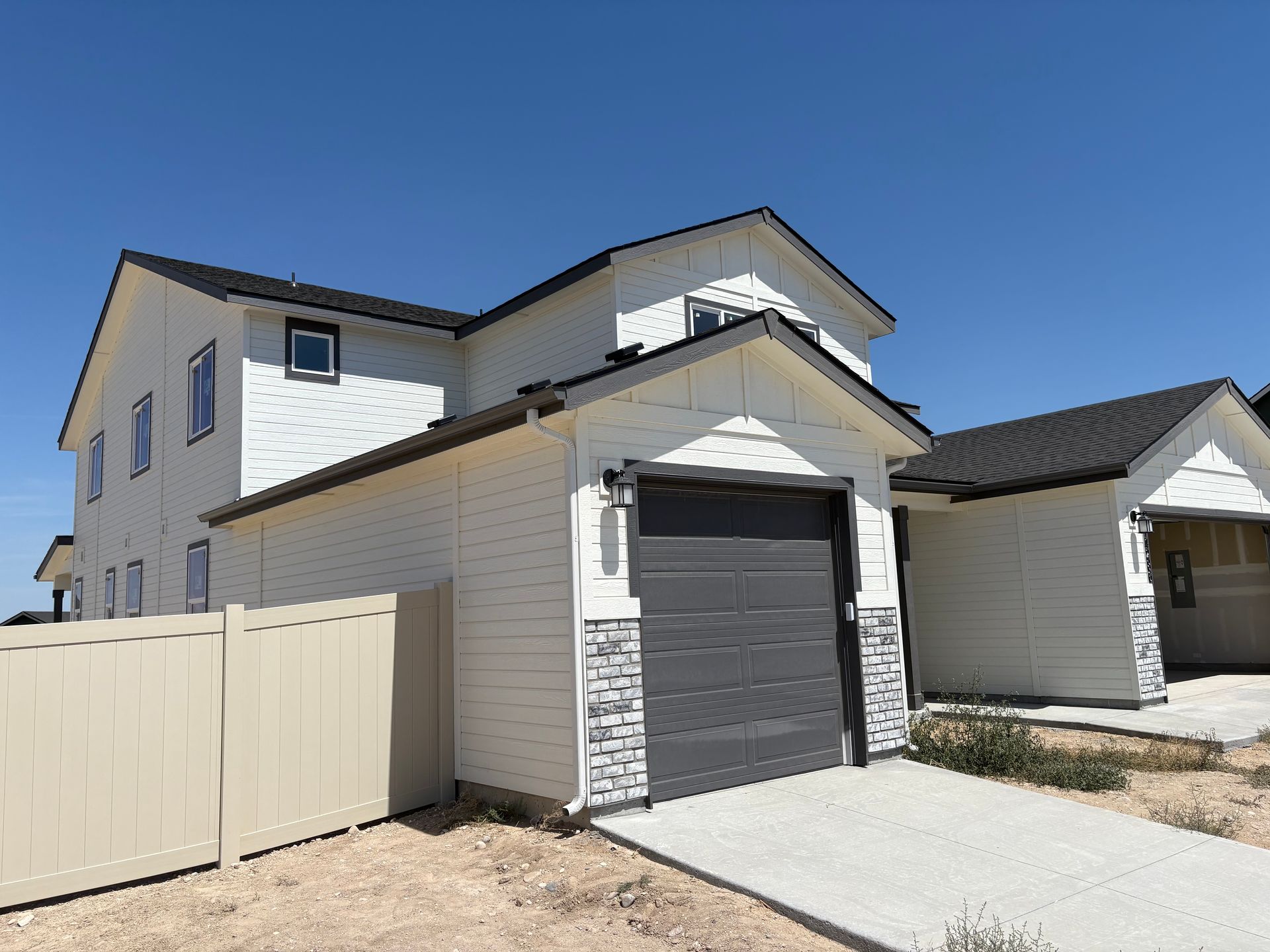 White two-story house with detached garage; gray garage door, black roof, beige fence, clear blue sky.