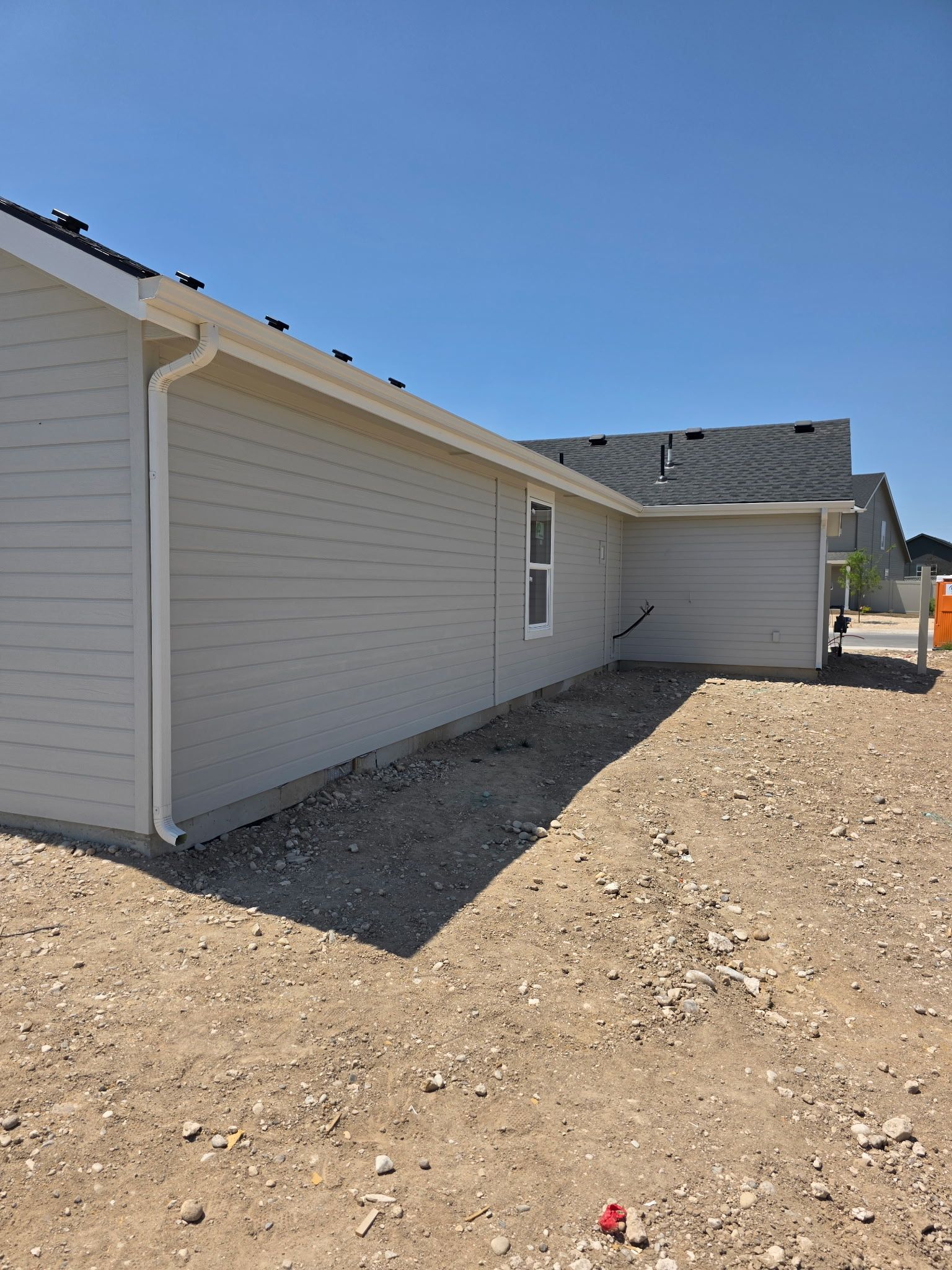Light gray house with white trim, gutters, and a single window sits on a gravel lot under a blue sky.