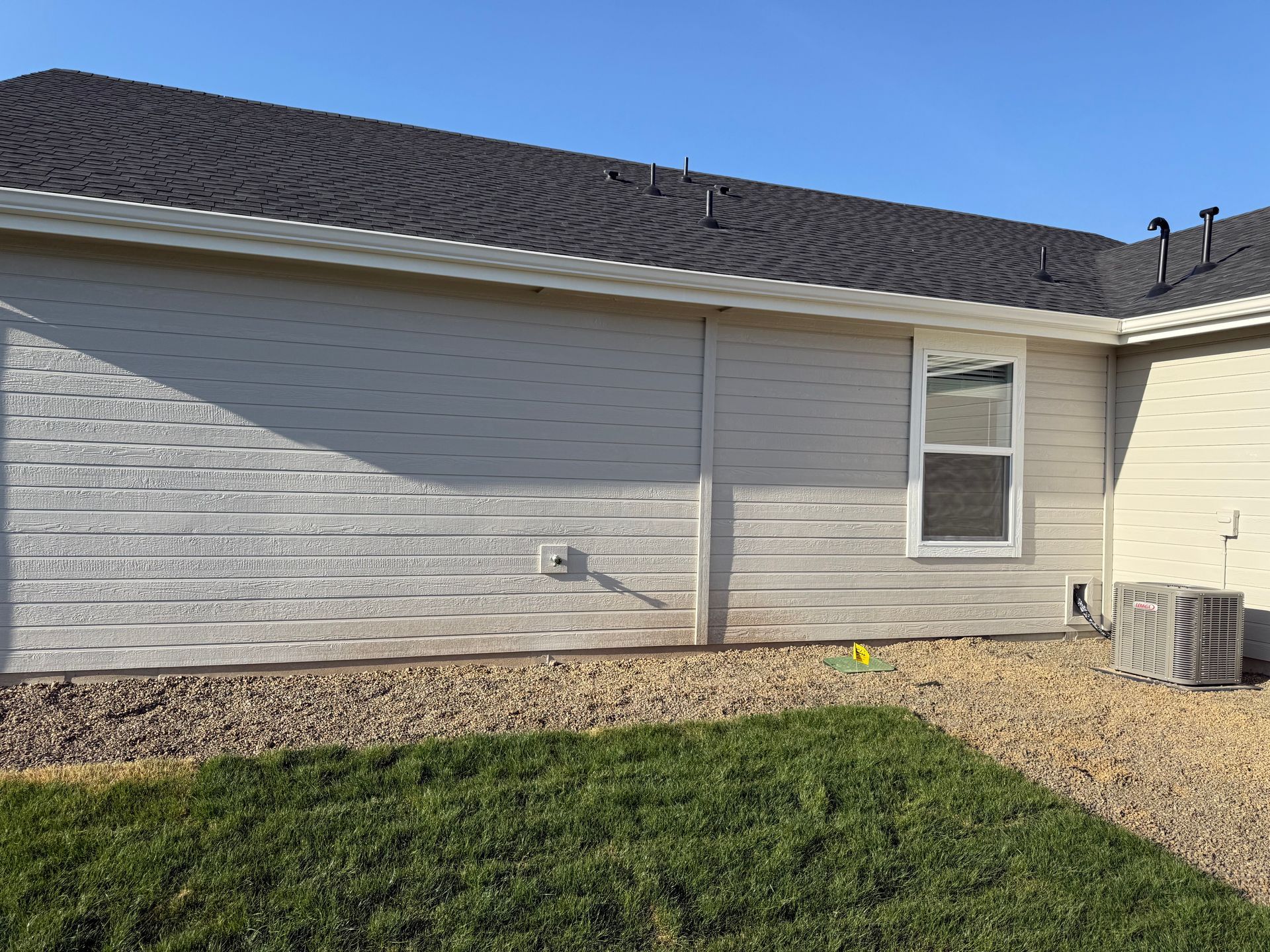 Exterior of a house with light siding, dark roof, window, and air conditioning unit on a gravel and grass yard.