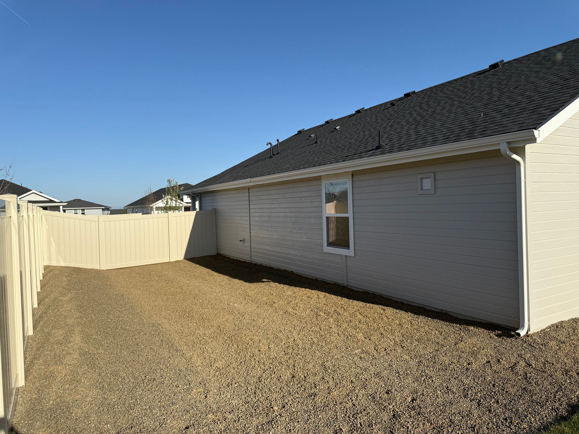 Beige siding and fence enclose a gravel-covered backyard with a window, against a clear blue sky.