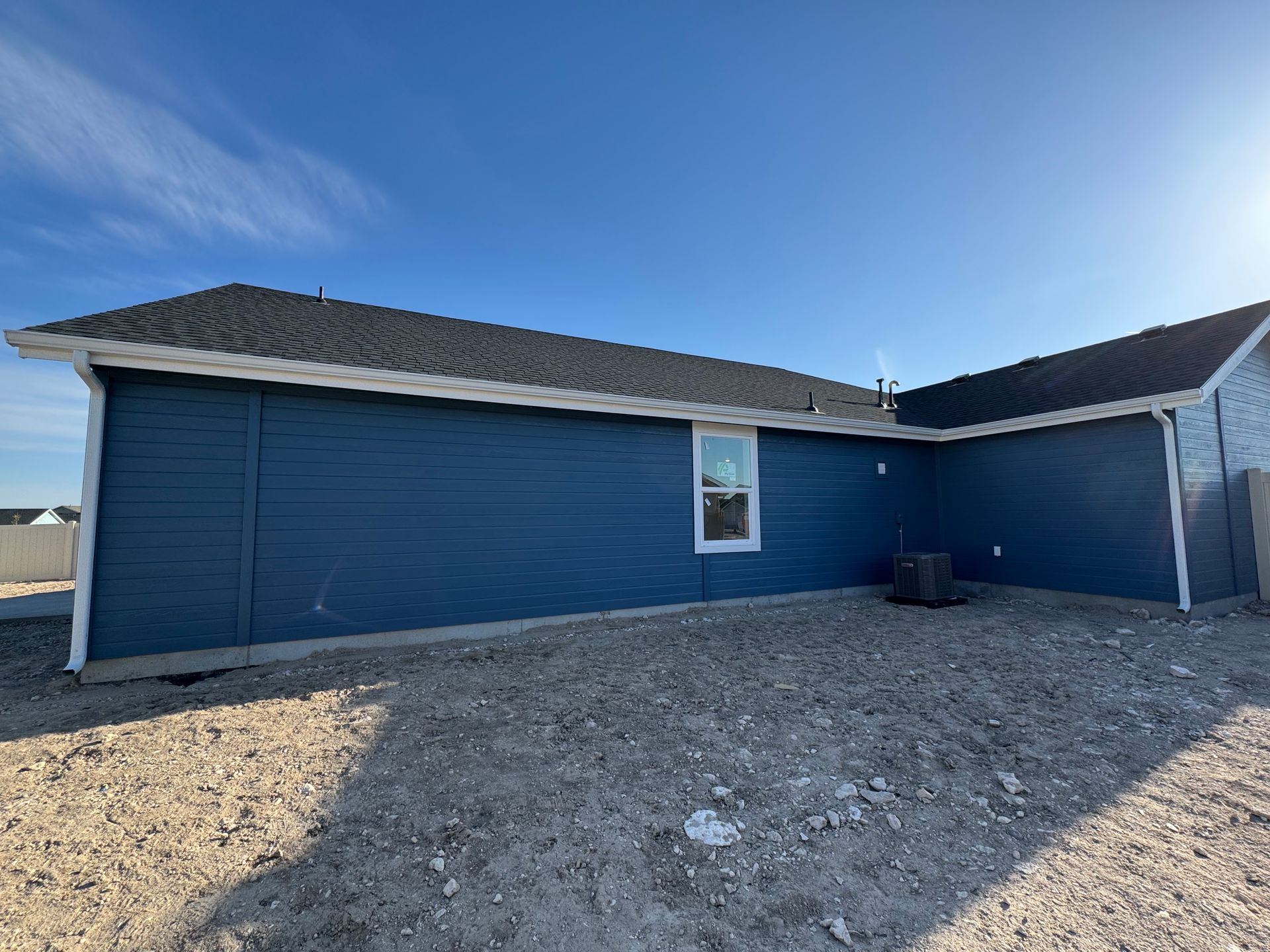 Blue-sided house with a dark roof and white trim under a clear, blue sky, set on a gravel lot.