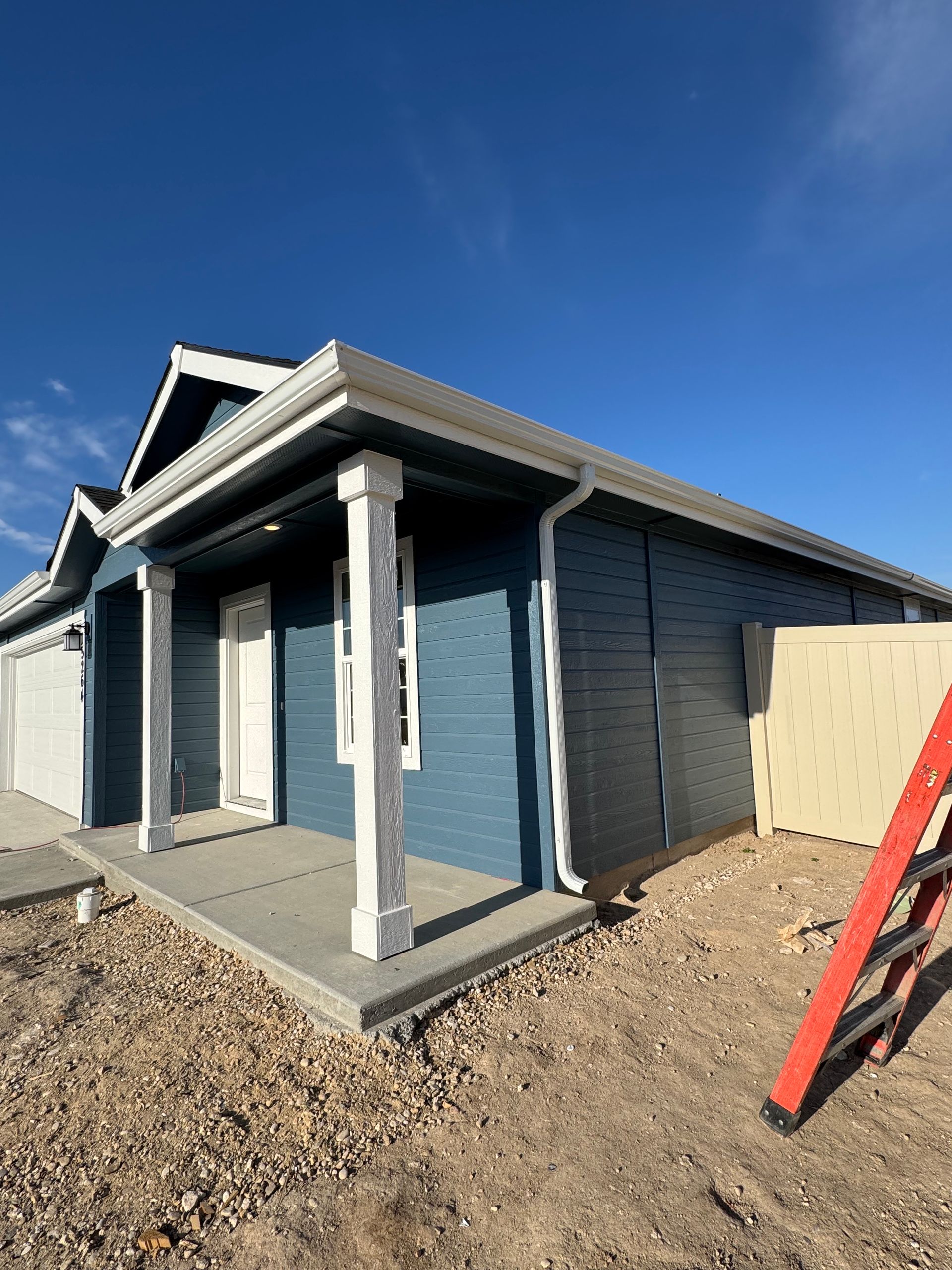 Blue house with white trim, porch, and a red ladder against a tan fence under a blue sky.