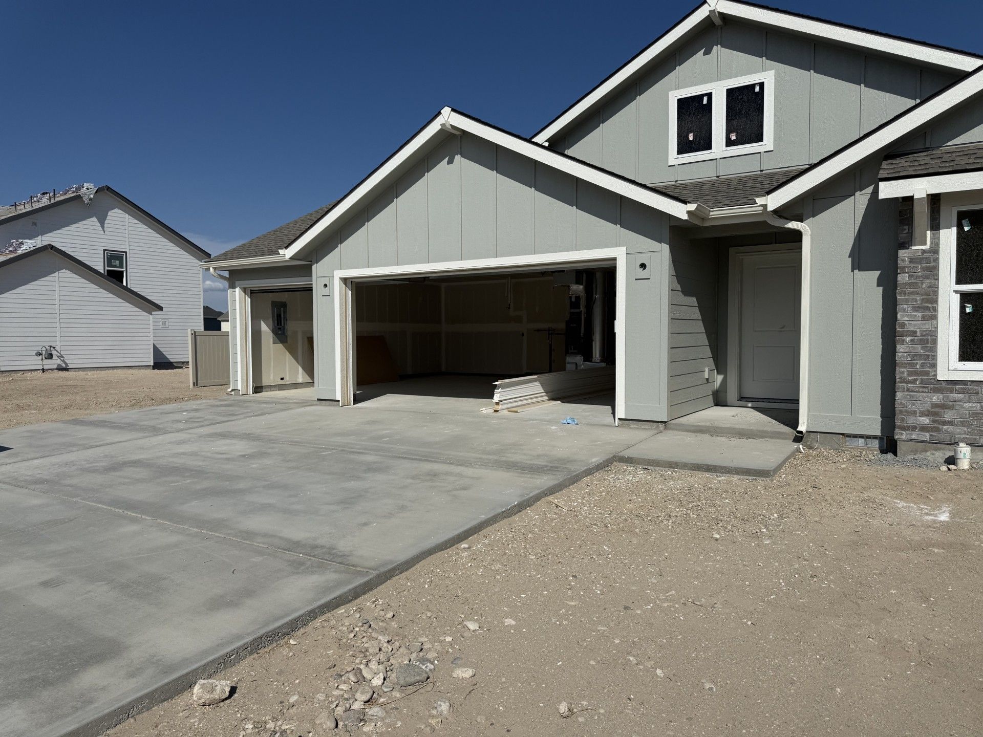 New house under construction, light gray siding, open garage, concrete driveway, gravel yard.