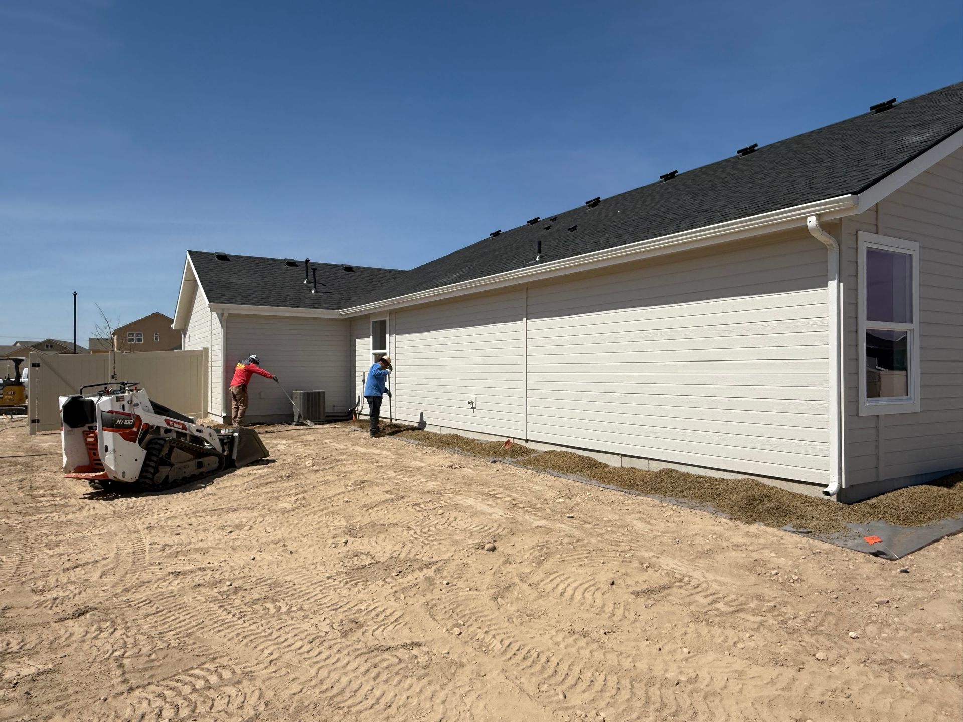 Two people working near a house under construction; dirt ground, blue sky.