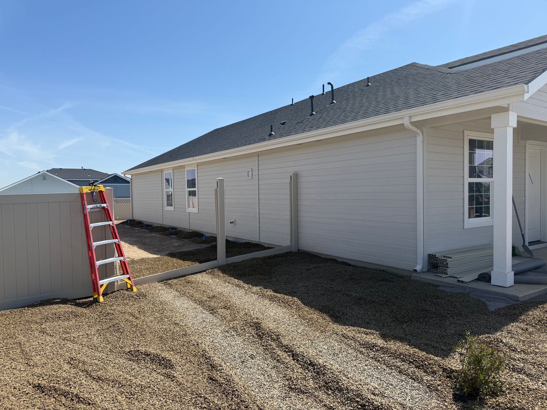 Side of a house with light siding, gravel, and a ladder against a utility structure; clear sky.