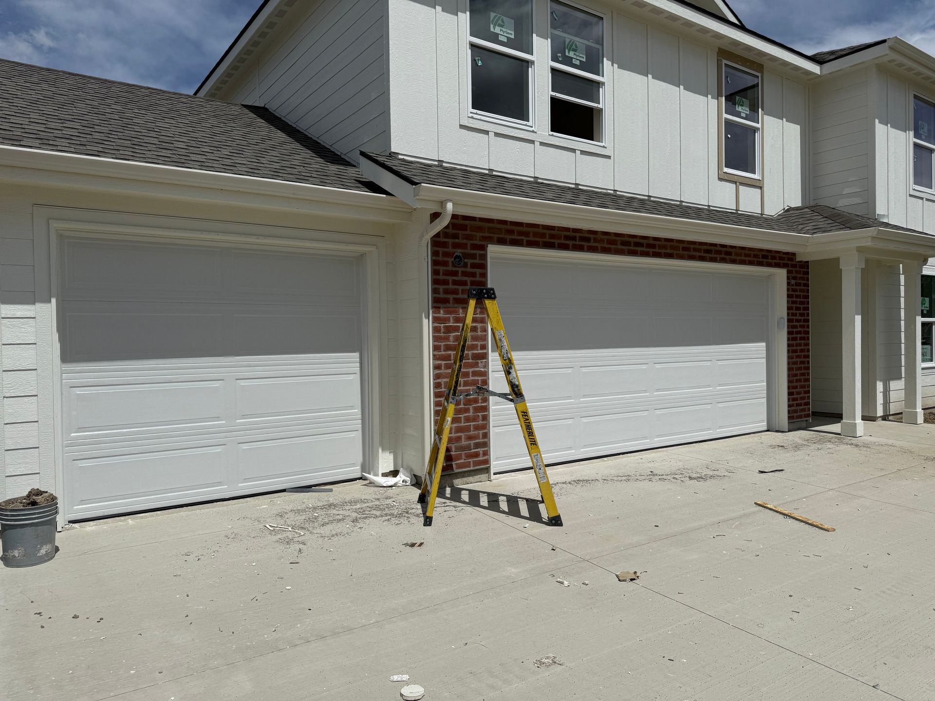 Two-story house with two white garage doors, a ladder, and a brick accent wall; construction site.
