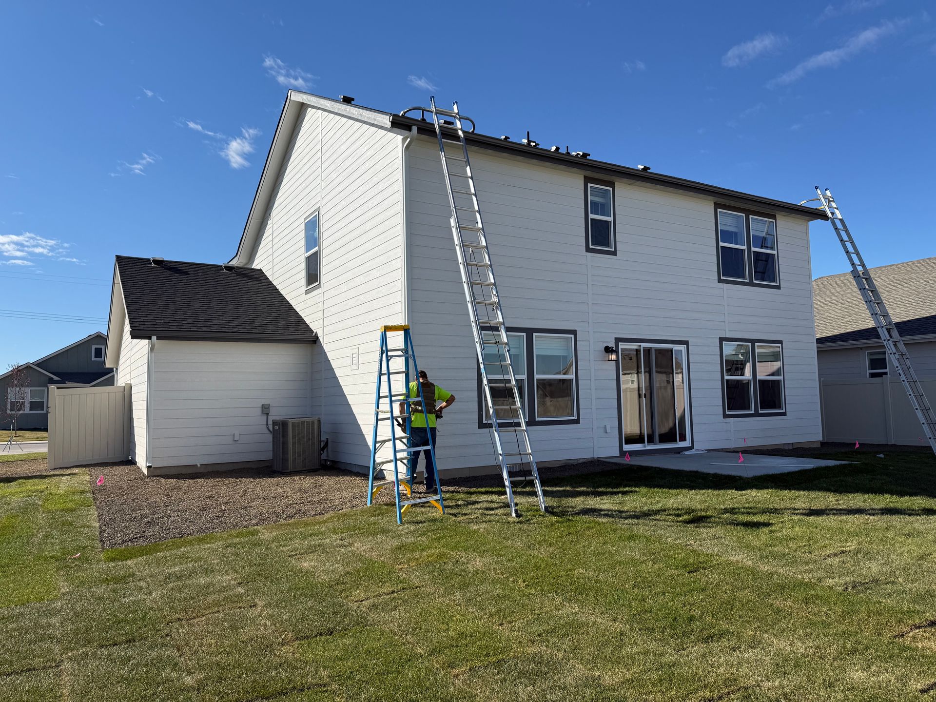 Person on a ladder, installing lights on a two-story white house with several ladders and clear blue sky.