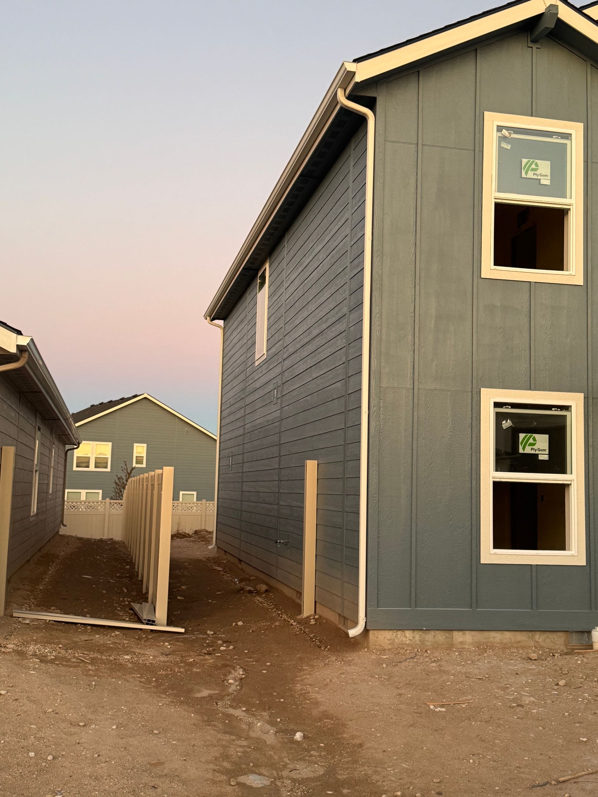 Side of a blue house with windows, next to a pathway and a wooden fence, under a colorful sky.