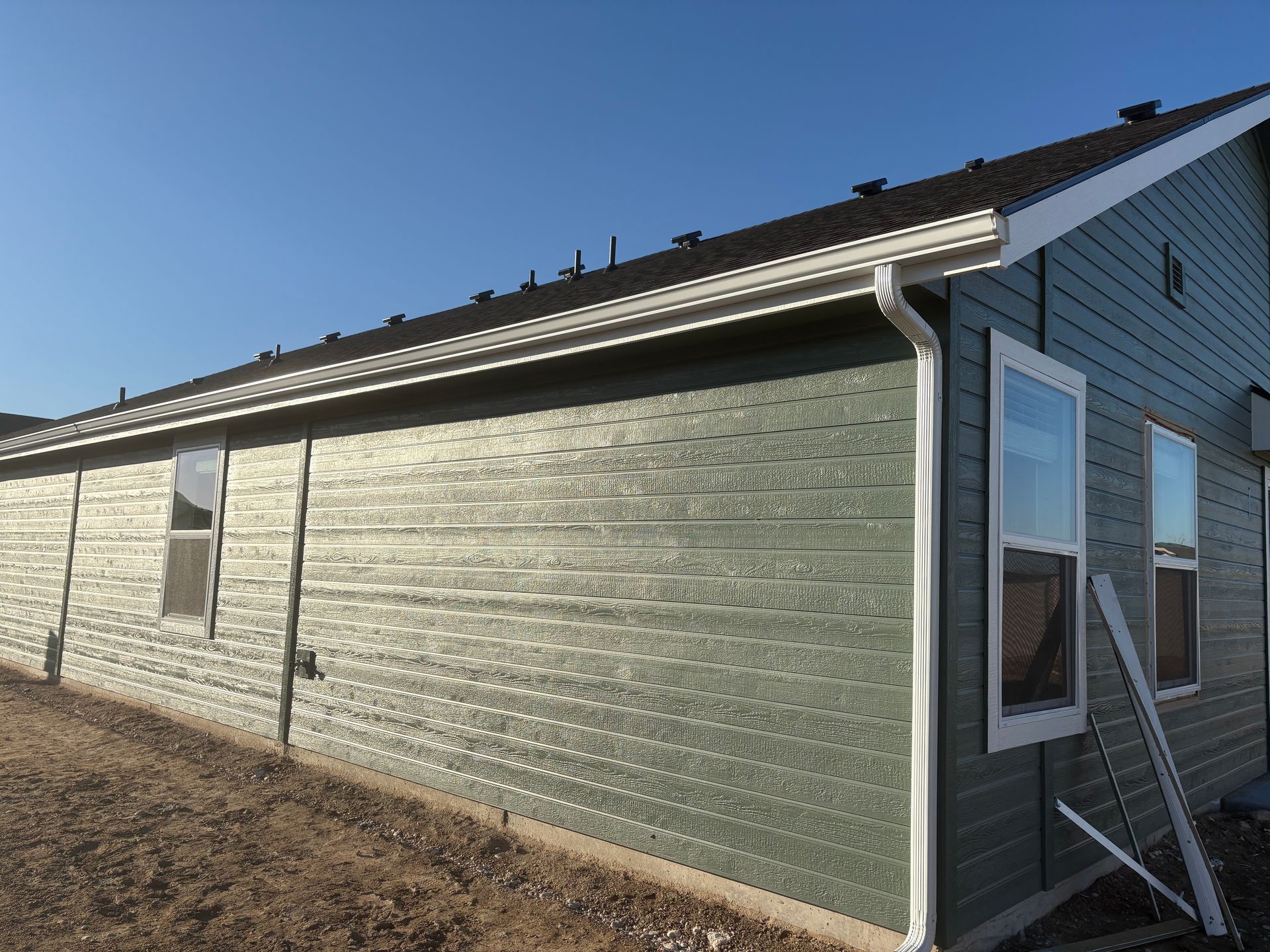 Green siding on a house with white trim, windows, and a roof against a blue sky.