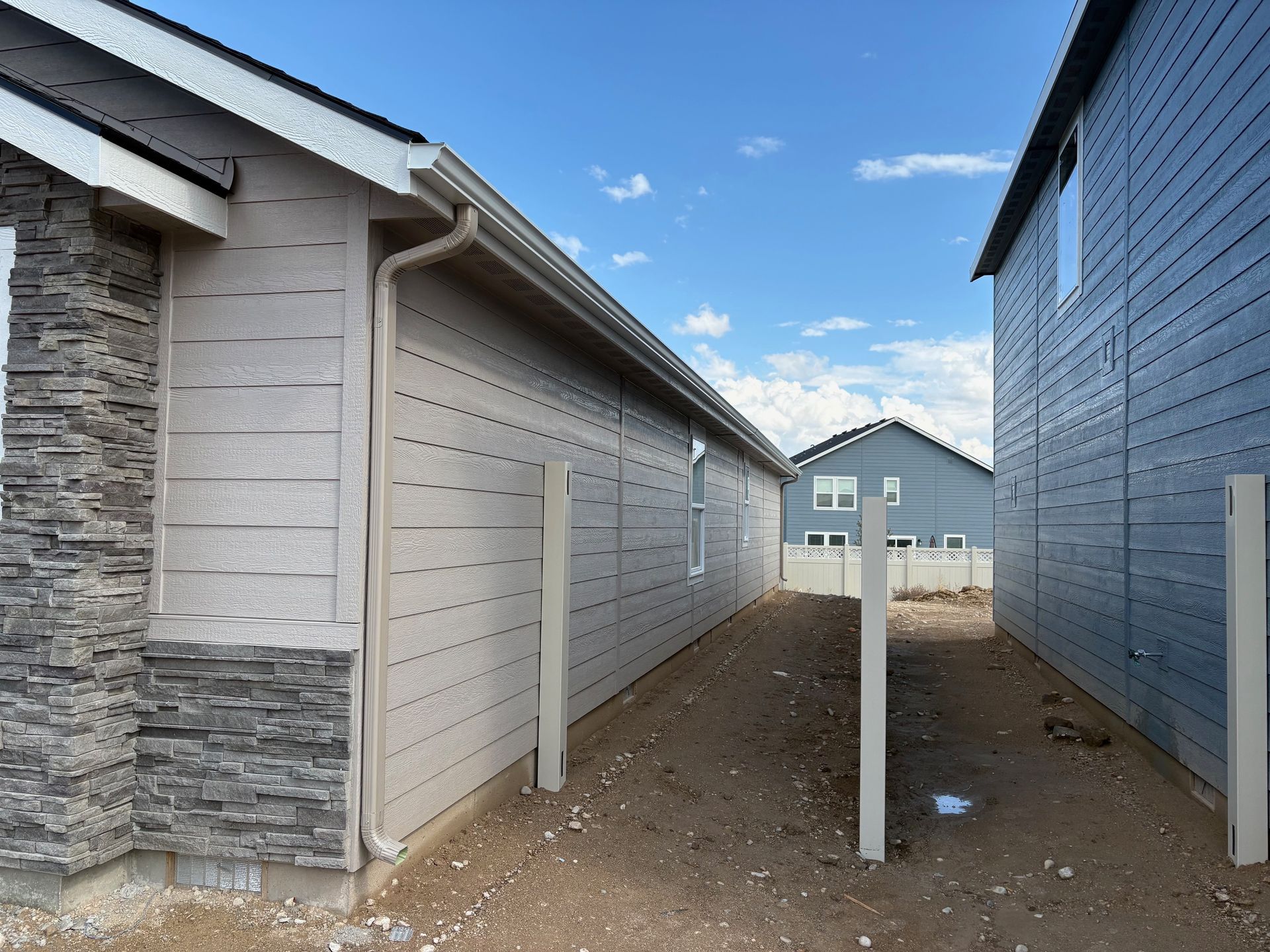 A narrow pathway between beige and blue houses, with a distant house in the center. Sunny day with a cloudy sky.