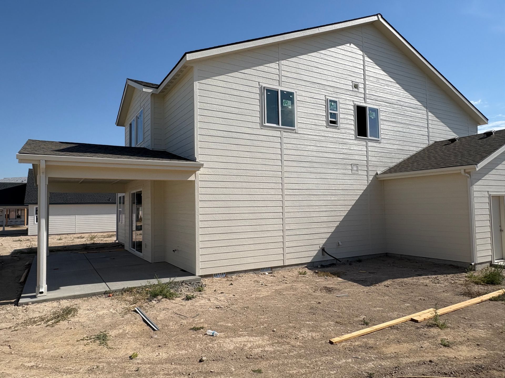 Two-story house under construction with light-colored siding, covered patio, and clear blue sky.