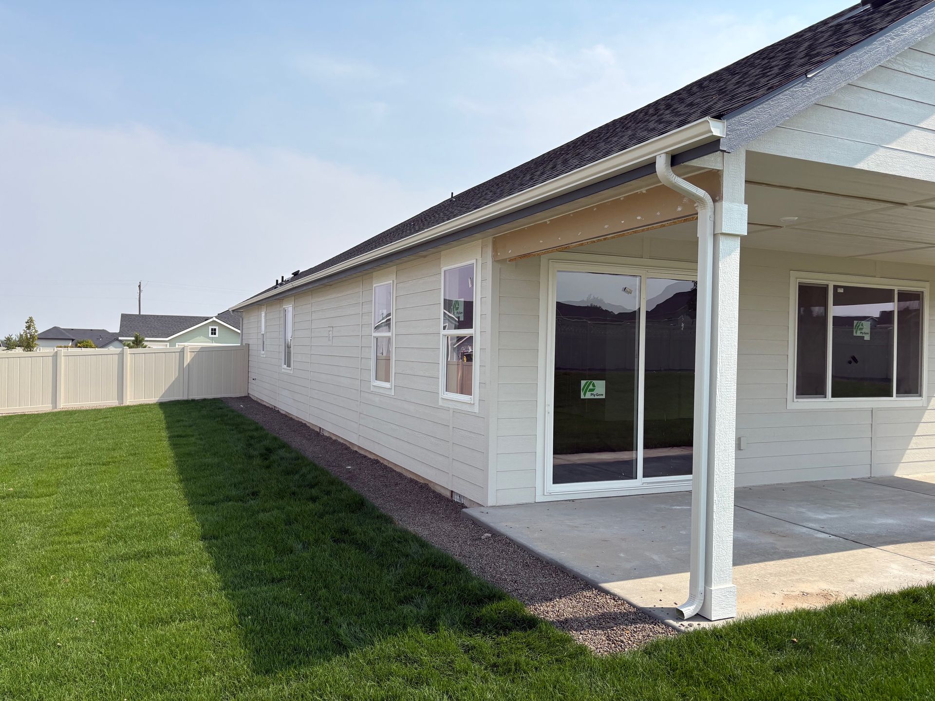 Backyard view of a beige house with a covered patio, green grass, and a white fence on a sunny day.
