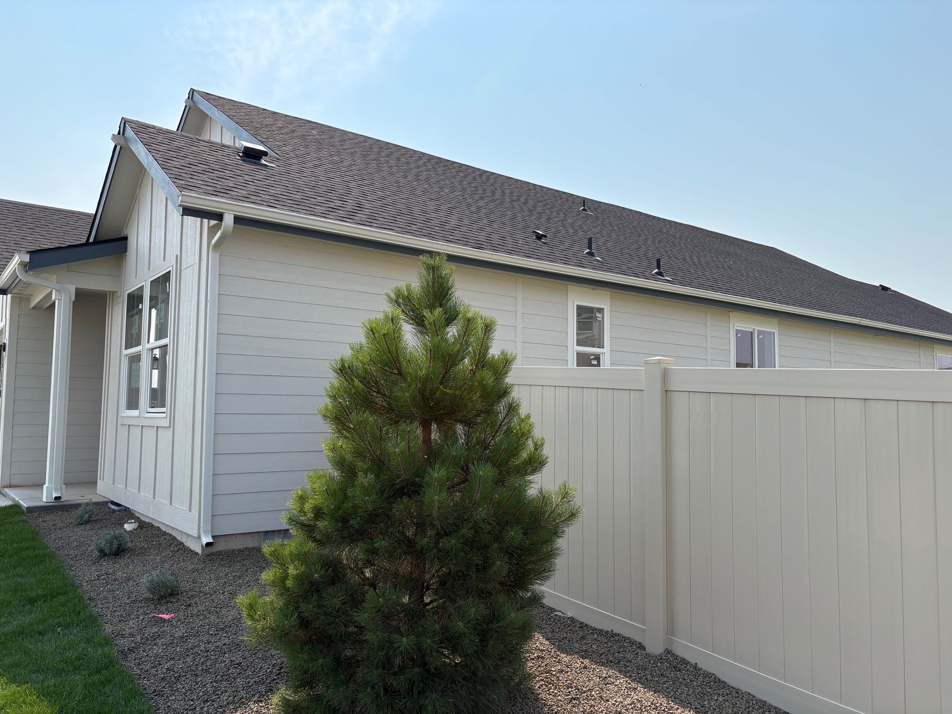 Light gray house with a dark roof and white trim, beside a beige fence and evergreen tree.