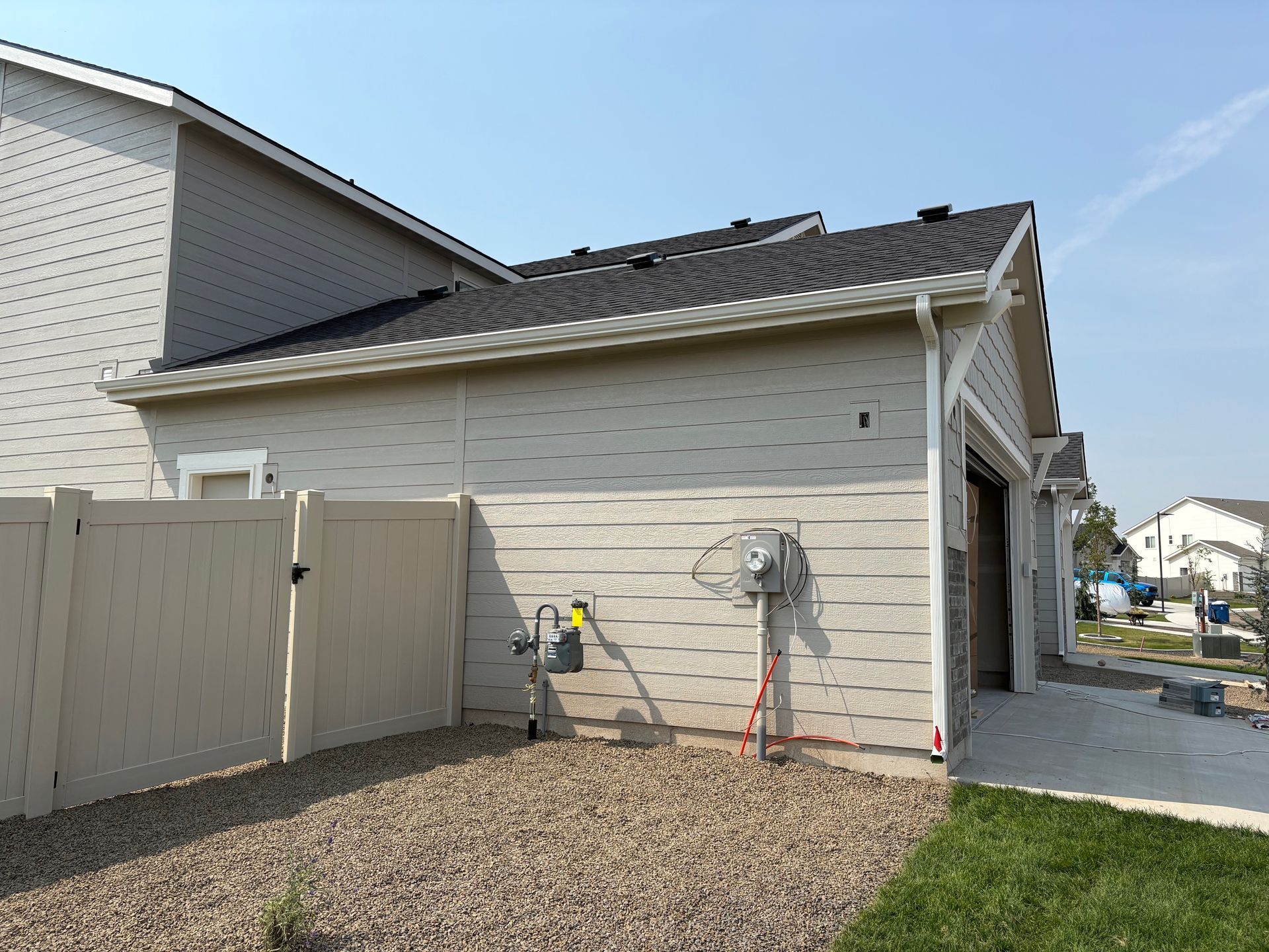 Side view of beige house with garage, beige fence, and gas meter on the wall. Gravel ground and blue sky.