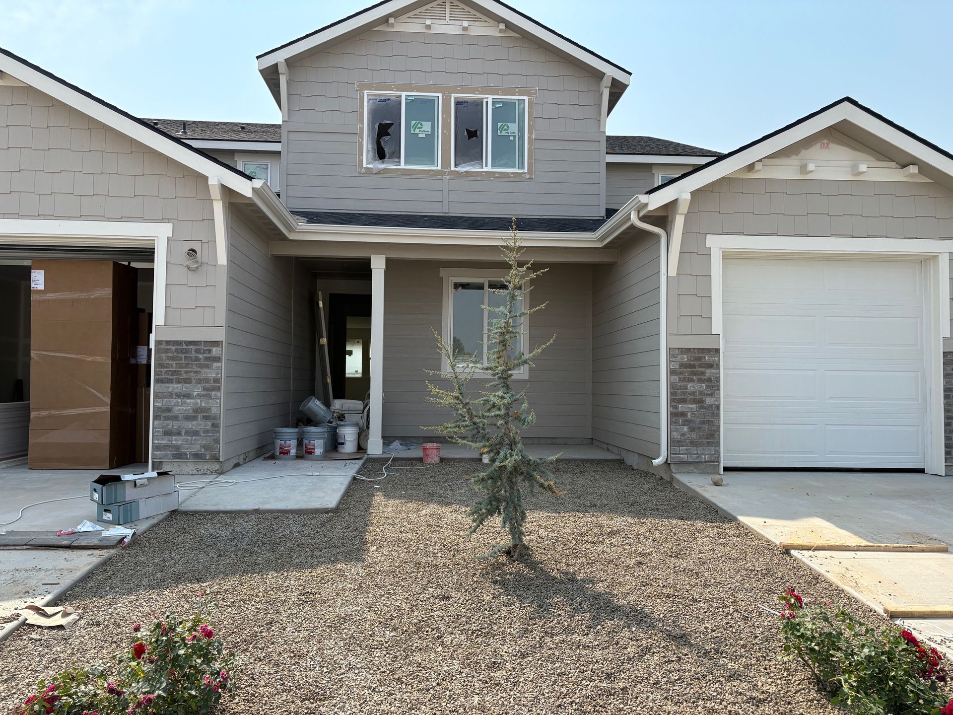 New house under construction, tan siding and stone facade, gravel landscaping, small tree.