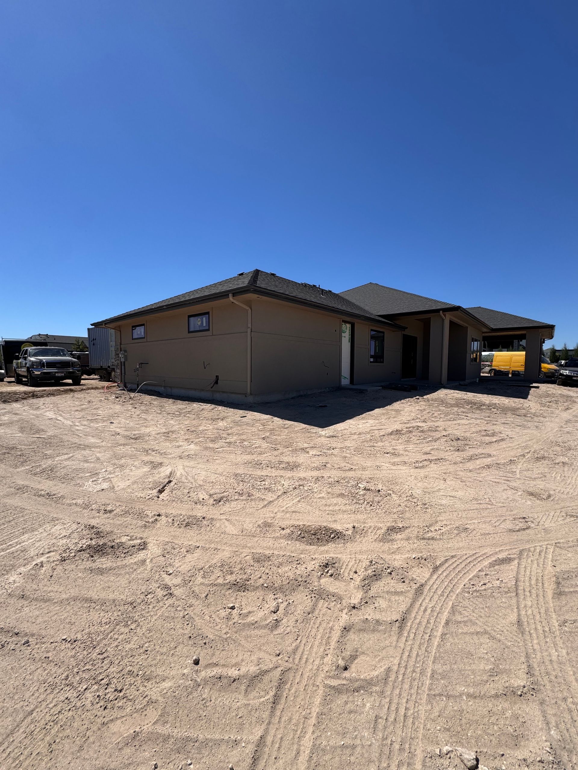 New house under construction on a dirt lot, gray roof, light brown exterior, blue sky.