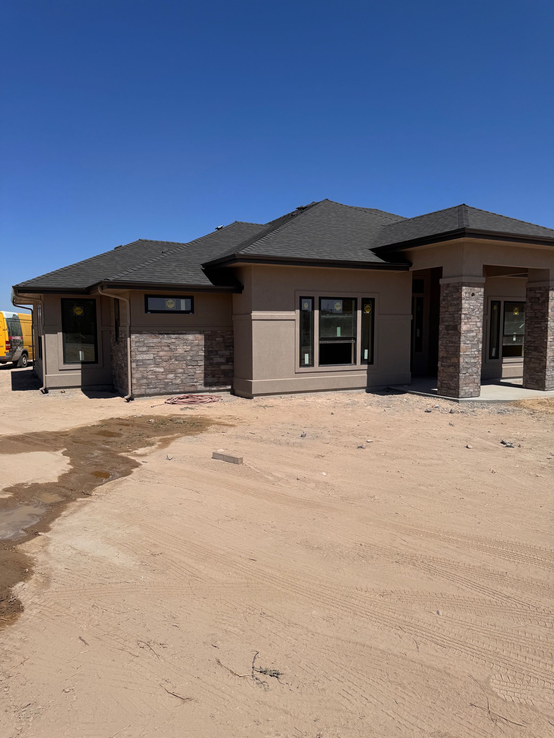 A single-story house under construction, featuring gray roof, tan stucco, and stone accents against a blue sky.