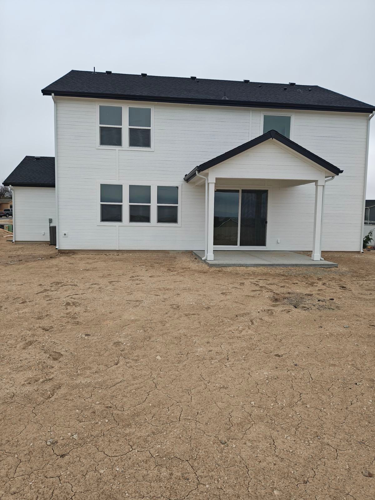 White two-story house with black roof, sliding glass door, and small porch on a brown dirt lot.