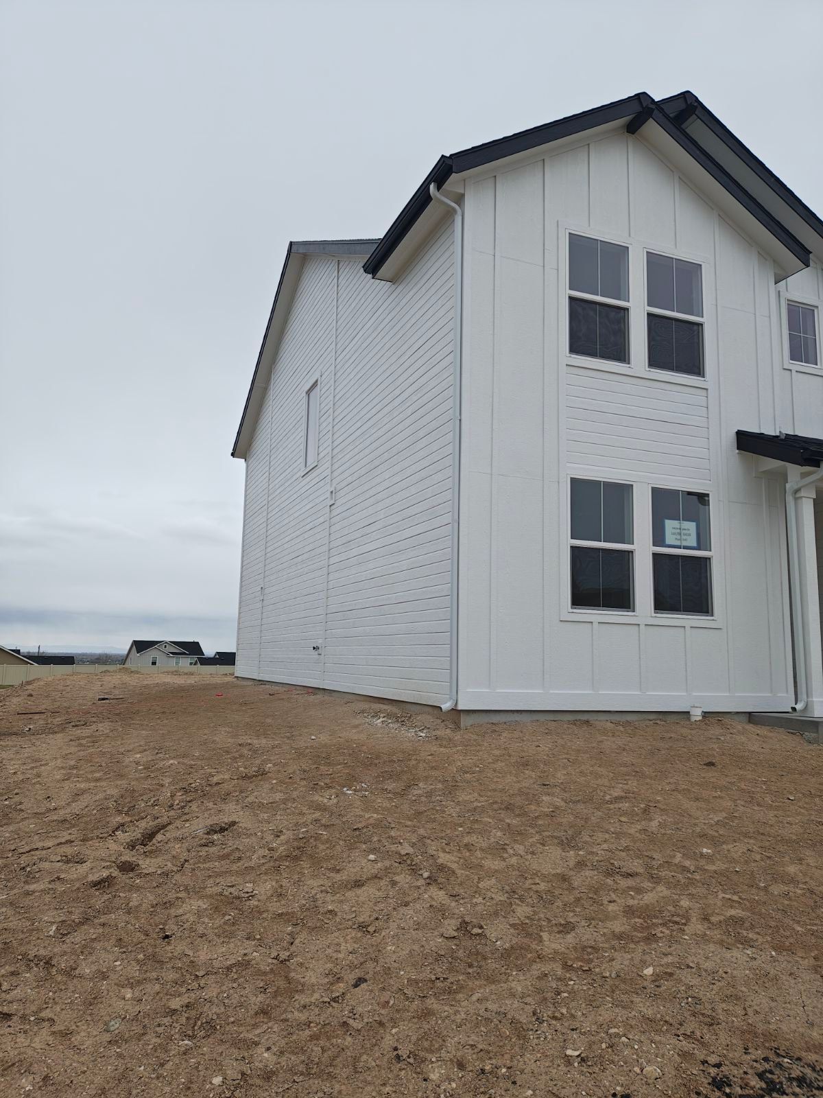 White two-story house under construction with vertical siding. Overcast sky, brown dirt in the foreground.