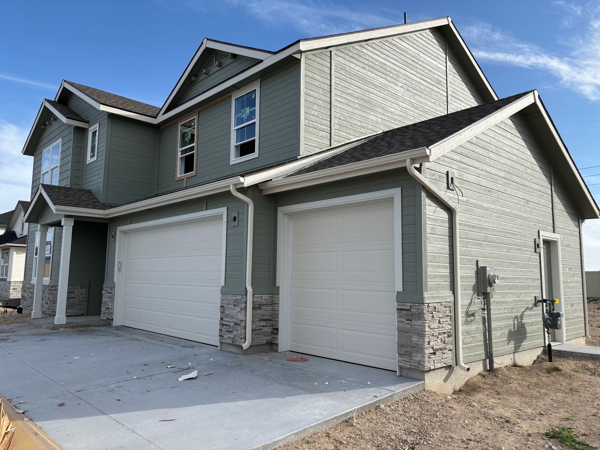 Two-story house with green siding, white garage doors, and stone accents under a blue sky.