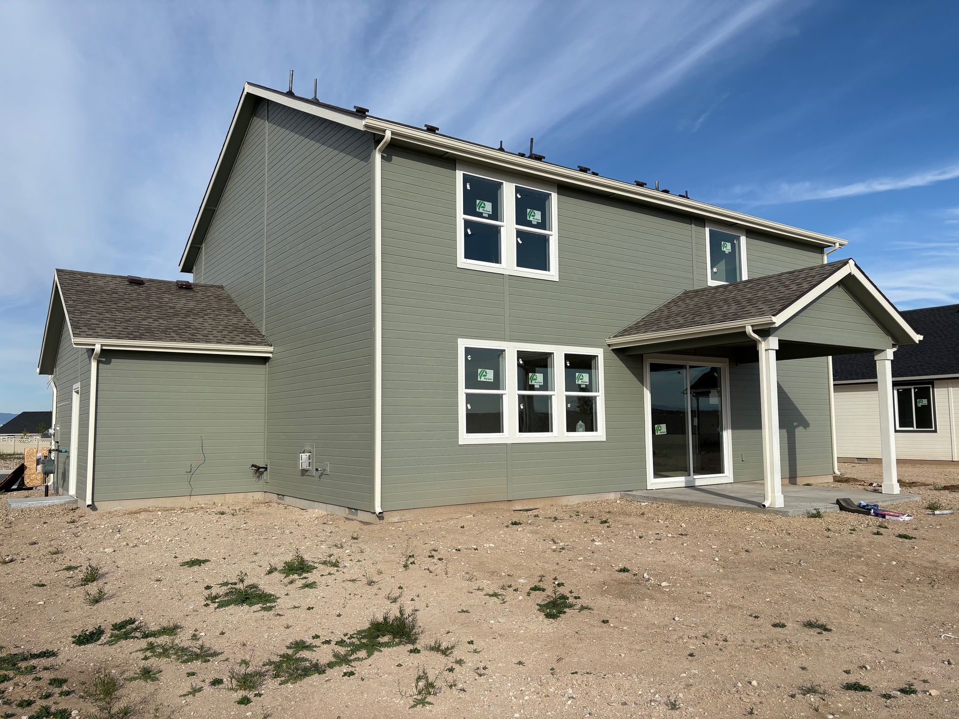 Newly constructed two-story house with green siding, white trim, and covered patio on a dirt lot under a blue sky.