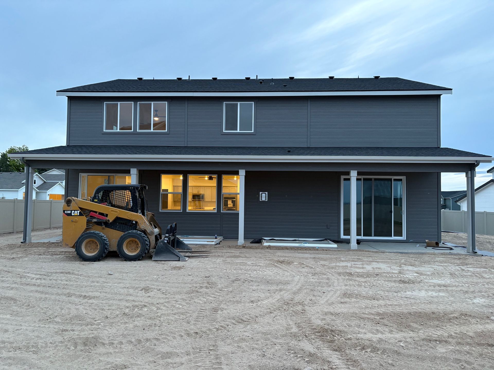 Back of a two-story gray house with a skid steer on a gravel lot.