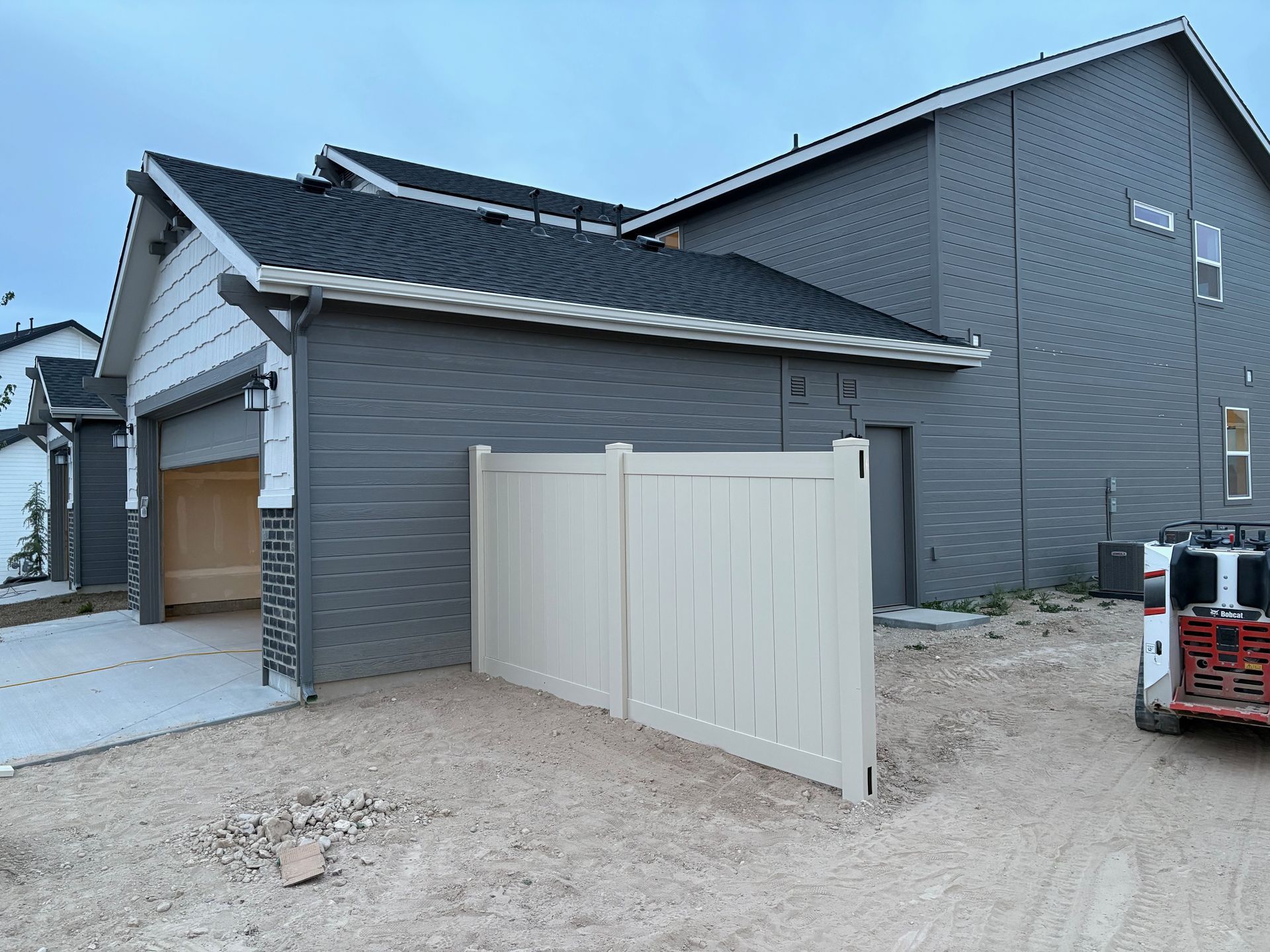 A two-panel cream vinyl fence in front of a gray-sided building with a garage and a dark roof.
