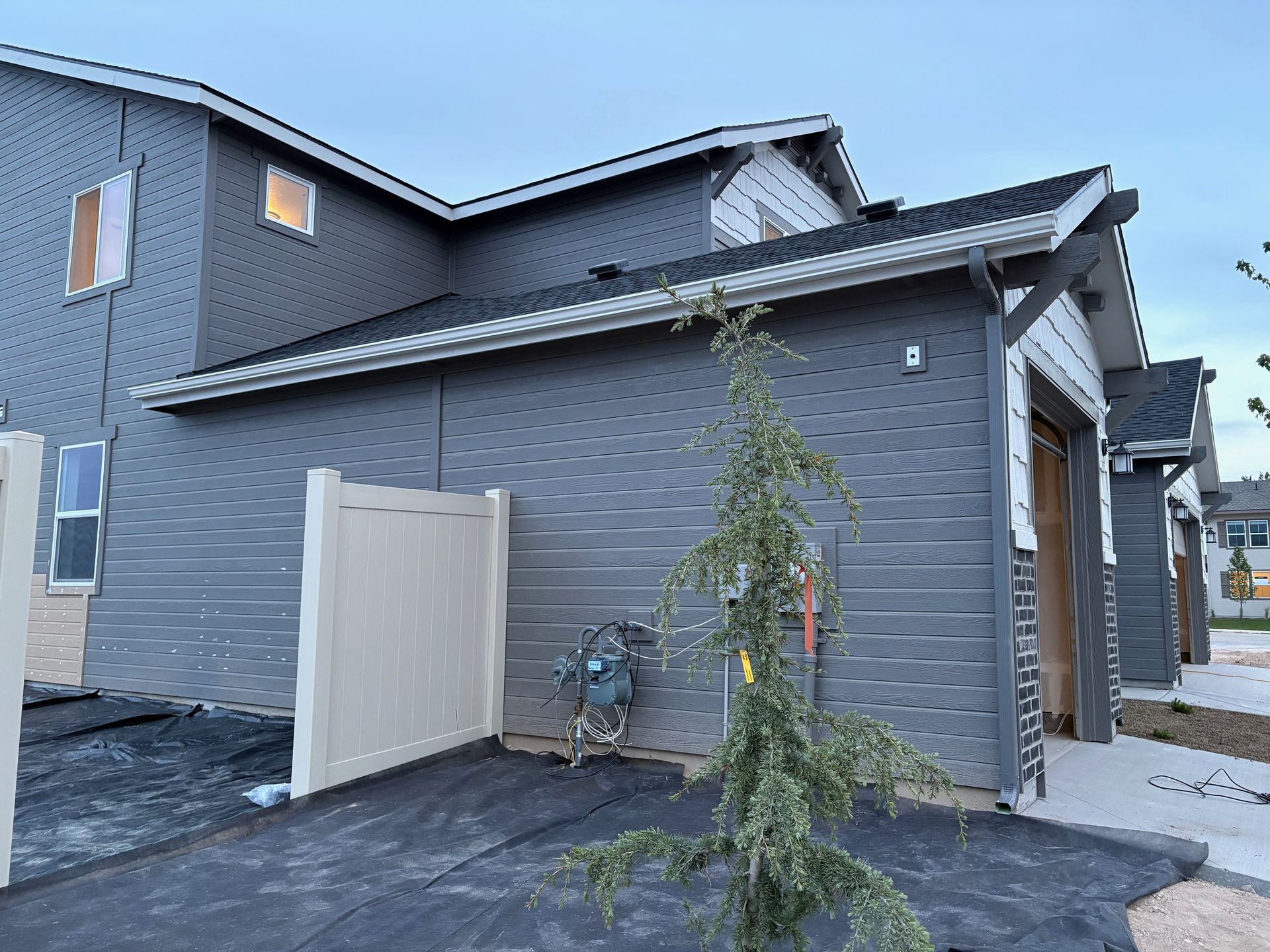 Gray siding of a house with a beige fence and a small tree in front.