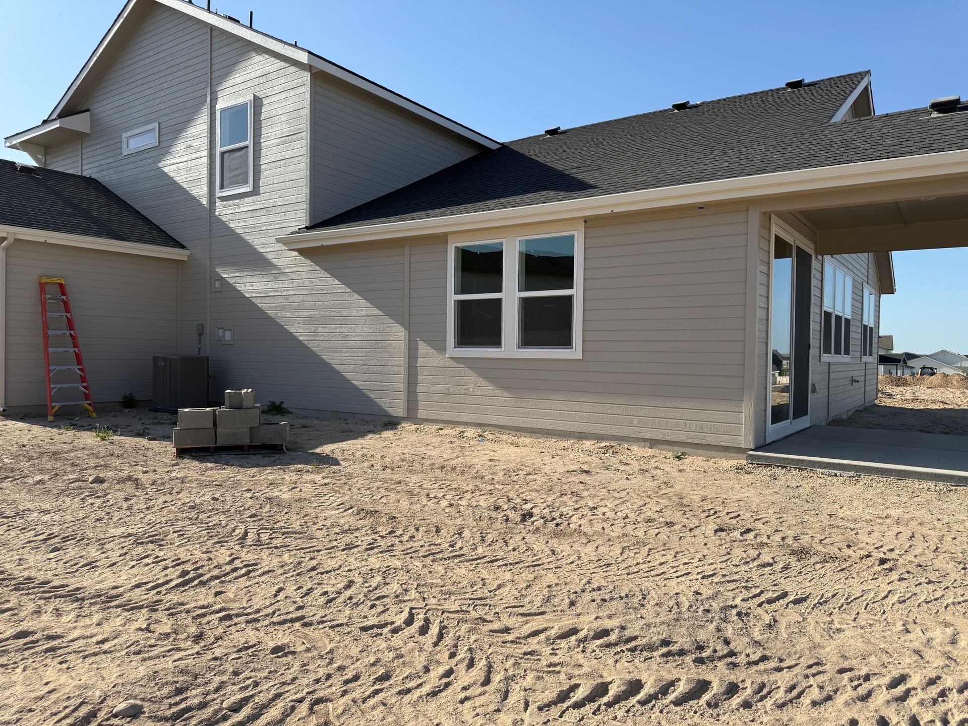 Tan house exterior with windows, roof, and a covered patio under construction.  Loose dirt and tire tracks in the foreground.