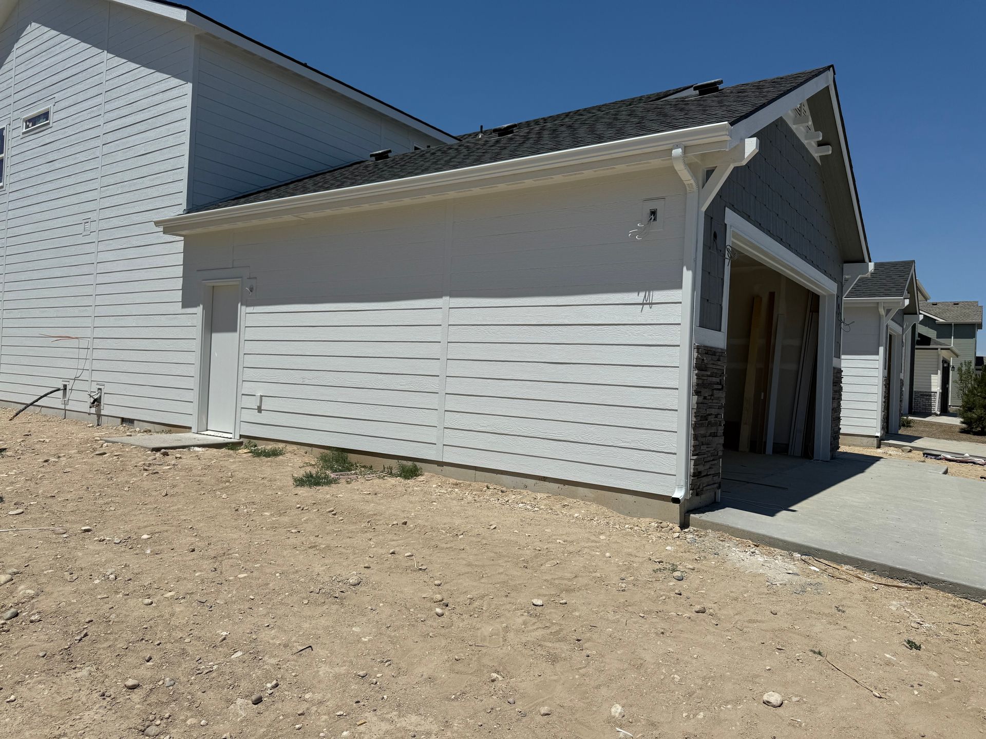 Light gray garage with dark roof, next to a larger gray house. Sandy ground in front.