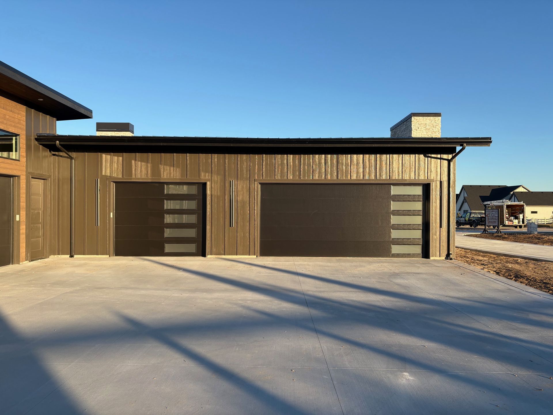 Two-car garage with brown siding and modern black garage doors; snow on the driveway under a clear blue sky.