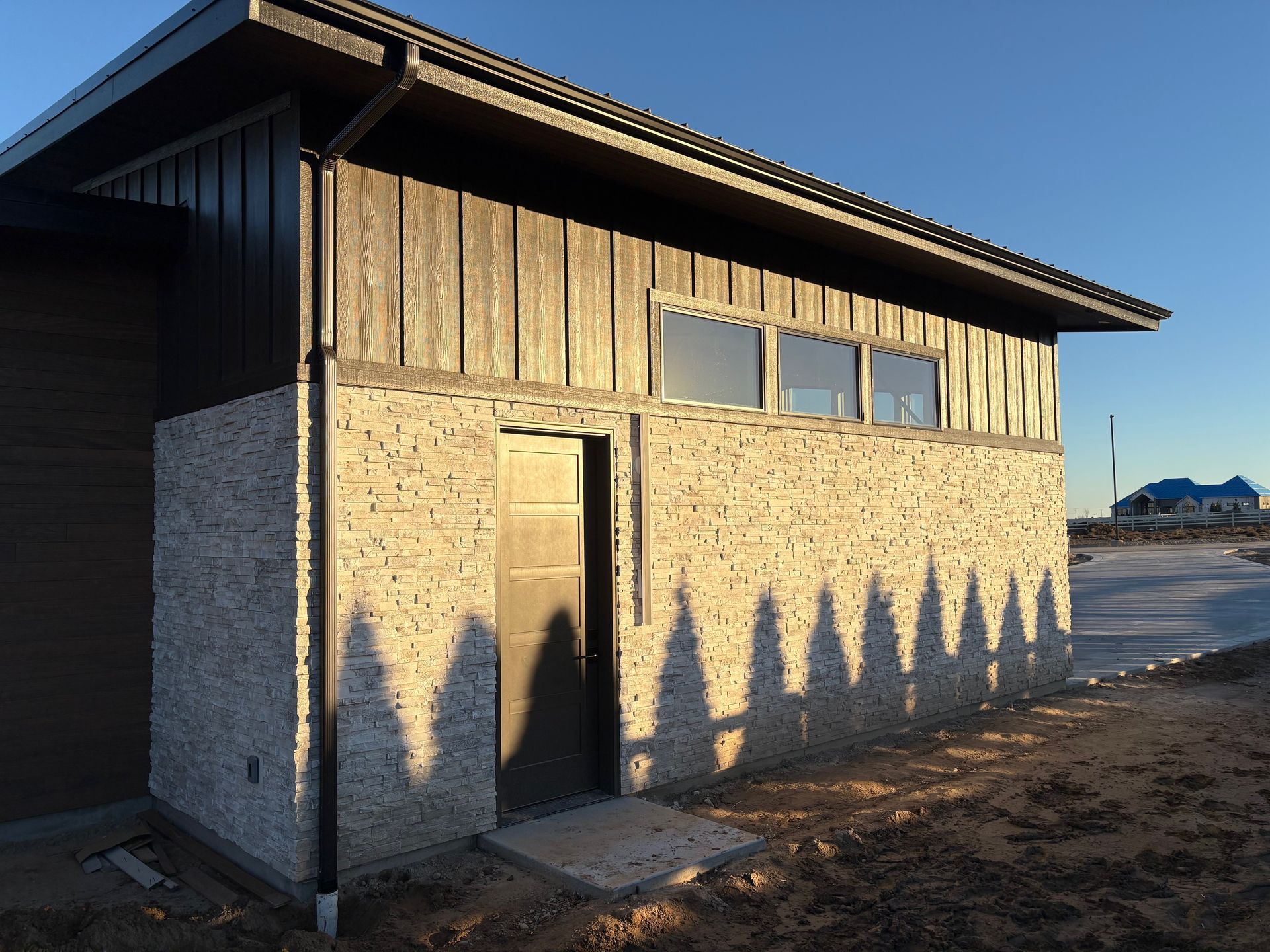 A building with wood siding and stone-like facade. Long shadows of trees cast on the building's side in sunlight.