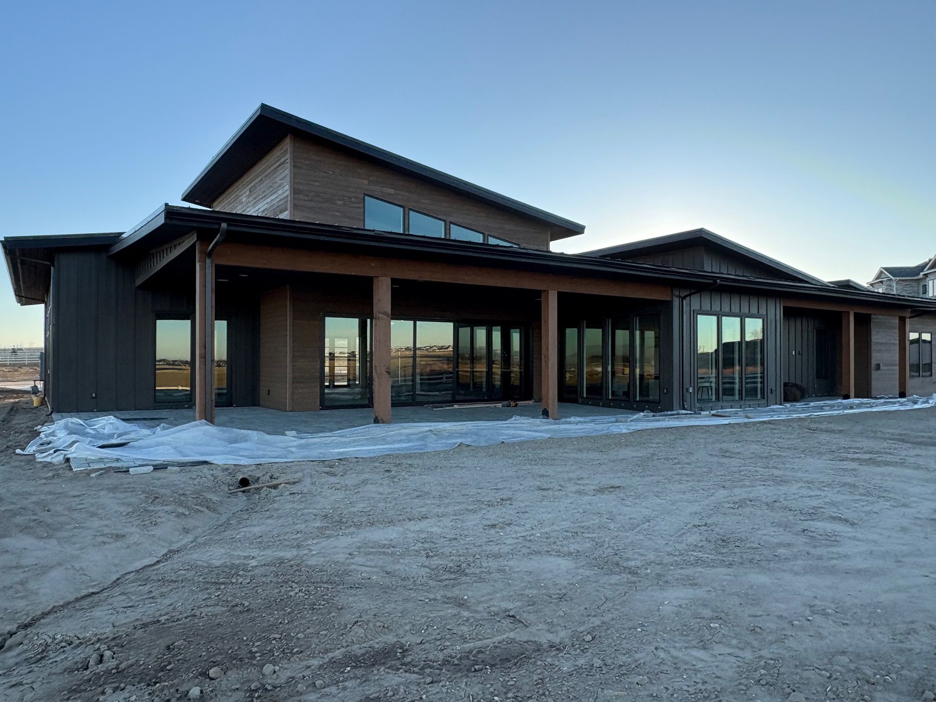Modern home exterior with large windows, covered porch, and brown siding on a gravel lot under a blue sky.